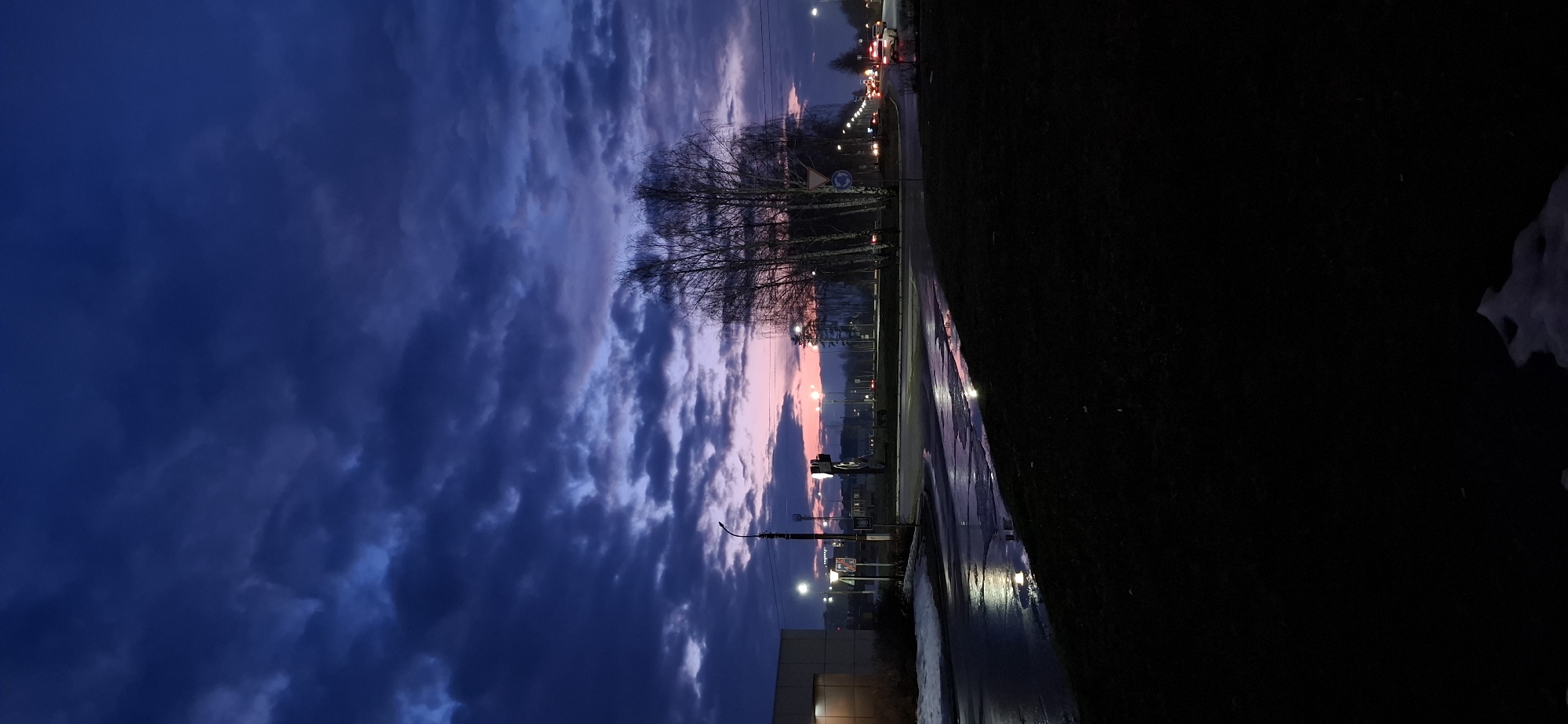 The image shows a twilight scene in what appears to be a city, possibly Chaikovsky, Russia. The sky is filled with dark, blue clouds with hints of pink and orange near the horizon. Bare trees stand along a wet roadside, reflecting light from street lamps and possibly car headlights. A building is visible on the left side. The pavement is wet, suggesting recent rain, and the grass is dark. Traffic lights and signs are visible in the distance. A patch of snow can be seen in the bottom right.