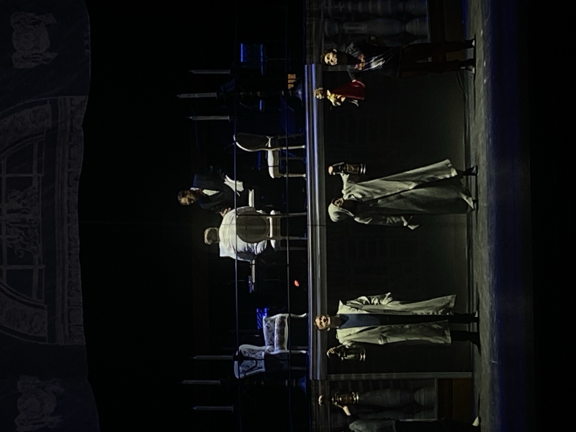 A stage production in Заволжье, Russia features actors in costume with props, including lanterns, chairs, and a backdrop resembling a building facade. A man at a piano, other standing actors, and set elements define the scene.