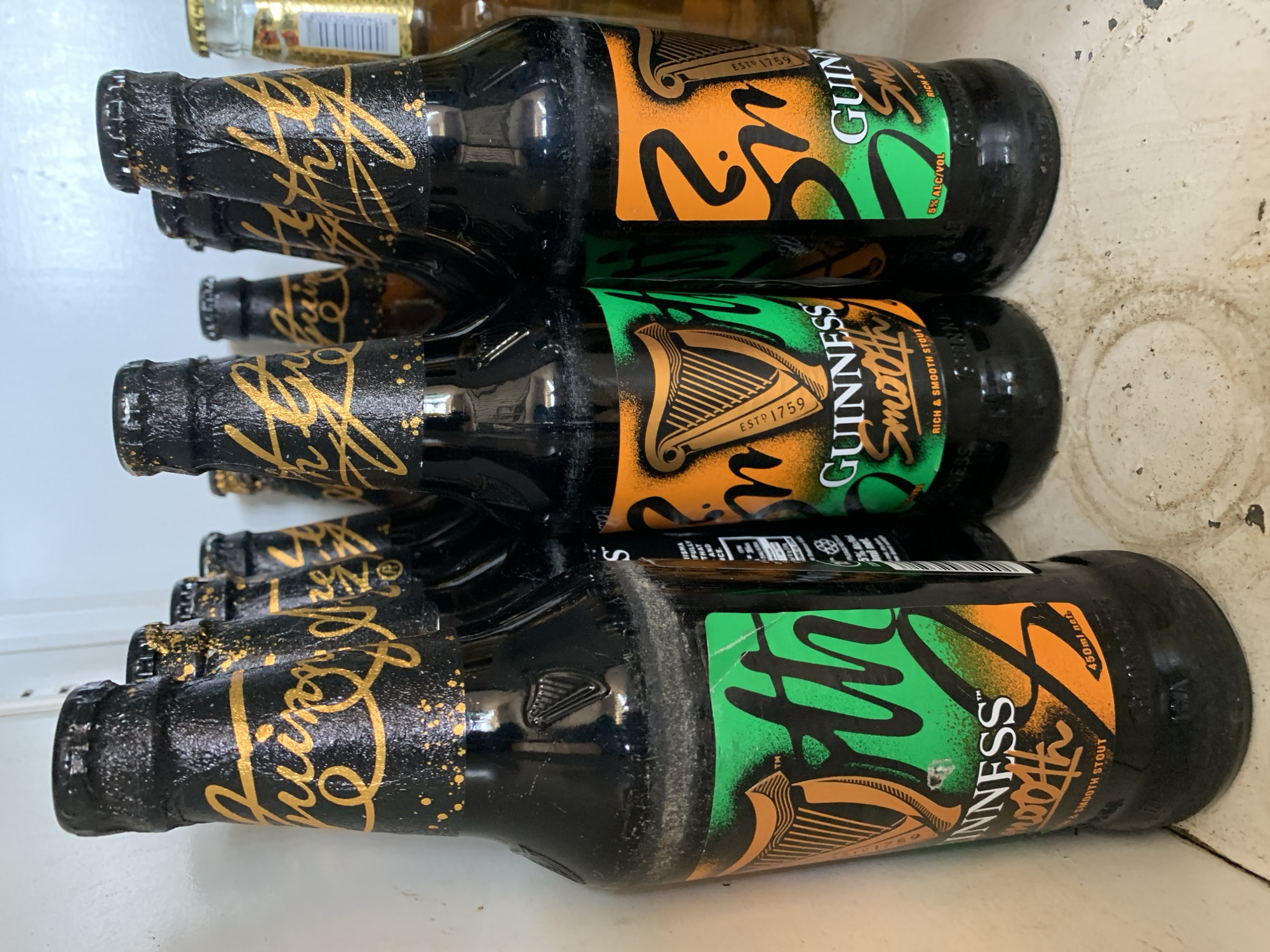 A stack of at least six bottles of Guinness Smooth stout are stacked horizontally against a white surface. The bottles are dark brown with a black neck. The labels feature the Guinness logo with a harp, and the words "Guinness Smooth" in a combination of green and orange colors. The necks of the bottles have golden decoration. Some of the bottles are dusty or have some residue on them. The background is a plain white, possibly a shelf or a refrigerator interior. There's another bottle partially visible above the stacked bottles. The location is not deducible from the image.