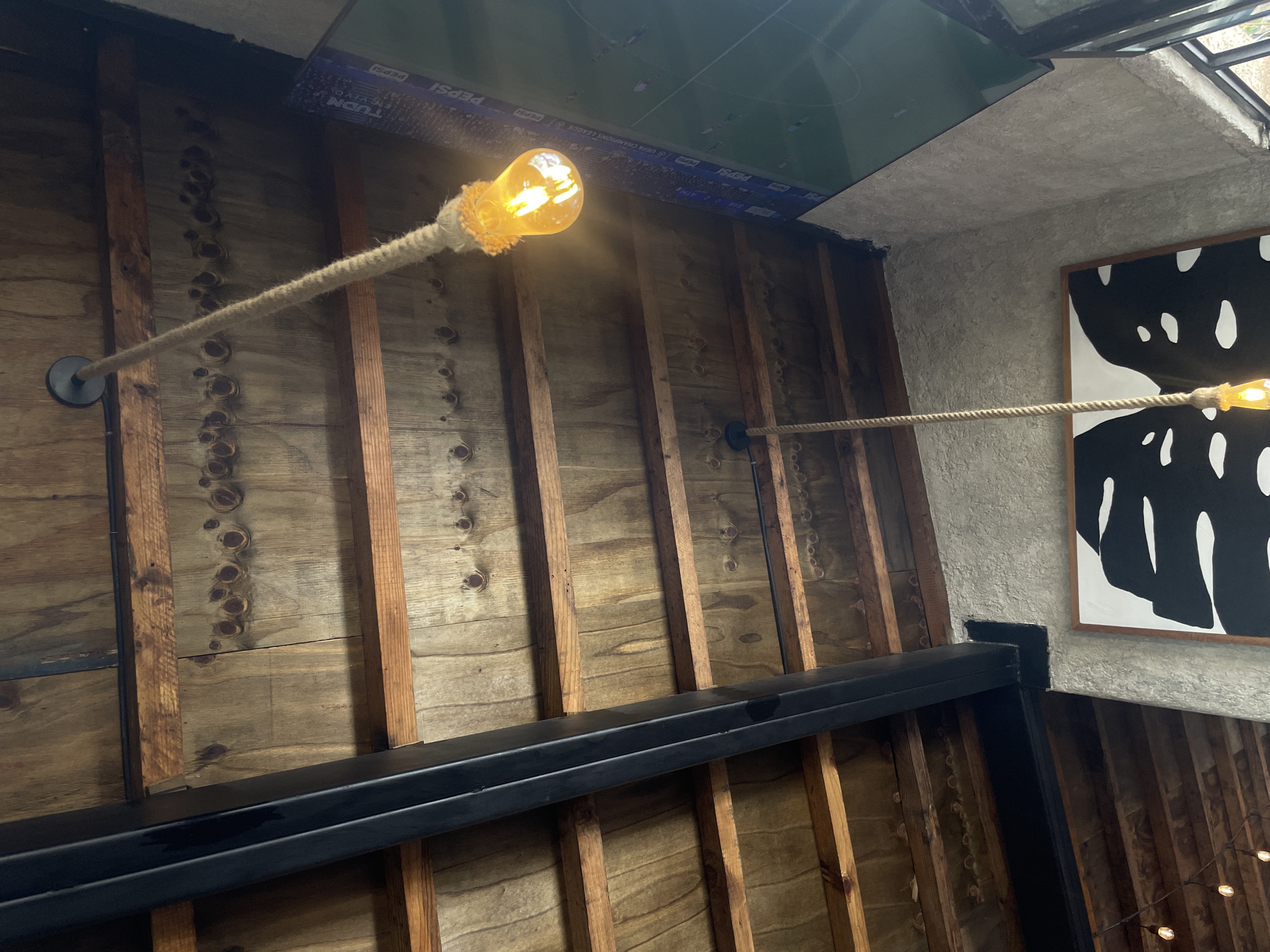 The image shows an interior space in Puebla, Mexico. The walls are made of rough, unfinished wood planks with vertical support beams. A thick, black steel beam runs horizontally across the lower portion of the frame. Two light fixtures, consisting of bare bulbs encased in rope netting, are mounted on the walls with metal fixtures. Above, a suspended ceiling displays a Pepsi advertisement and a sports broadcast. A black and white piece of art can be seen on the right wall.