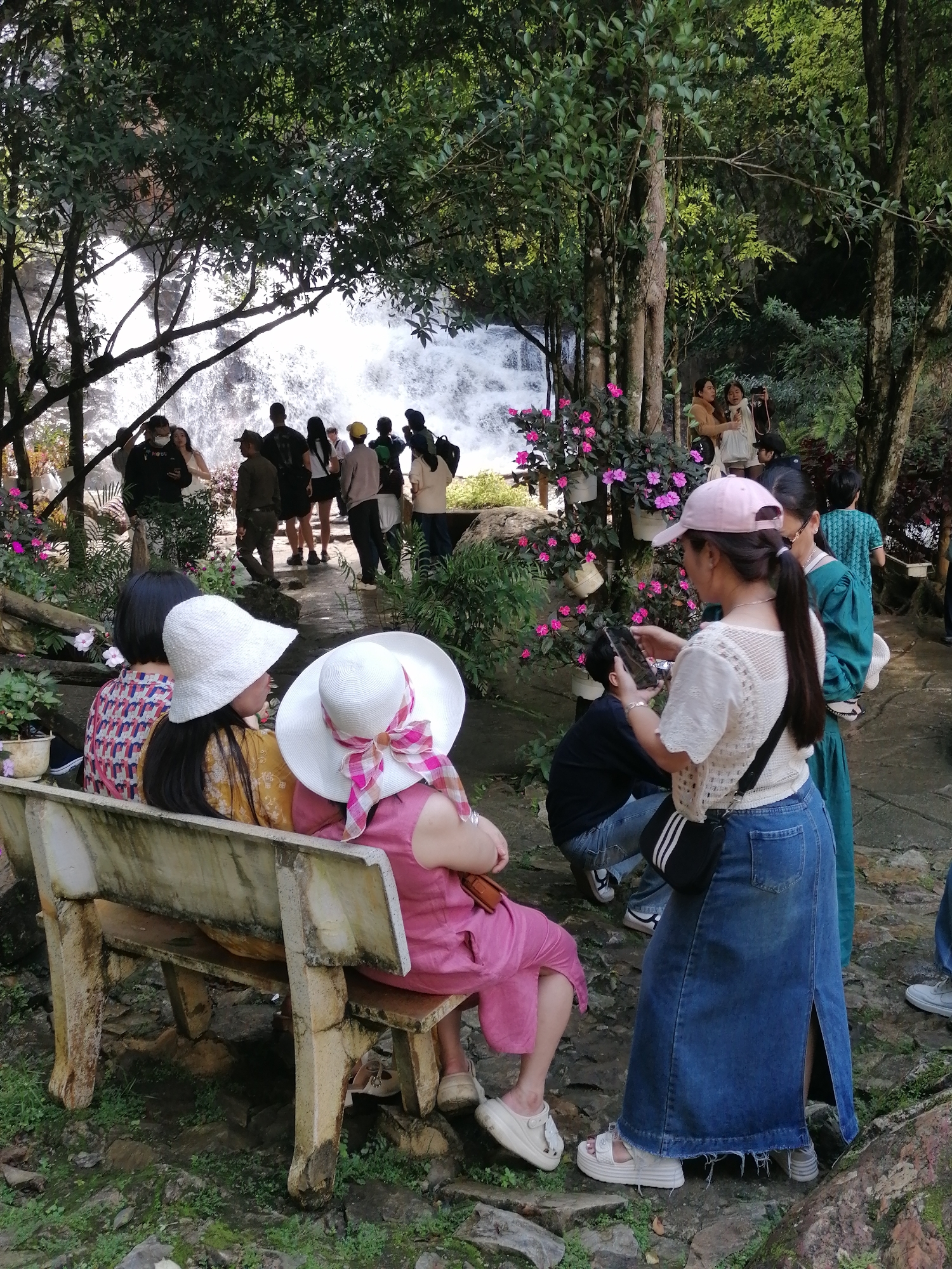 A group of tourists are gathered at Datanla Waterfall in Da Lat, Vietnam. In the foreground, two women sit on a weathered concrete bench, both wearing wide-brimmed hats. The woman on the right is wearing a pink jumpsuit and a white hat adorned with a pink and white checkered ribbon. The woman on the left is wearing a patterned top and a white bucket hat. Behind them, a woman in a white lacy top and a denim maxi skirt is holding a smartphone, seemingly taking a photo or video of a young man in jeans who is kneeling. To the right of this interaction, another woman in a teal dress and a young person in a green t-shirt are also present. In the background, a waterfall cascades down rocks, surrounded by lush green foliage and trees. Other tourists are visible on a path near the waterfall. The overall scene depicts a popular tourist destination with people enjoying the natural scenery.
