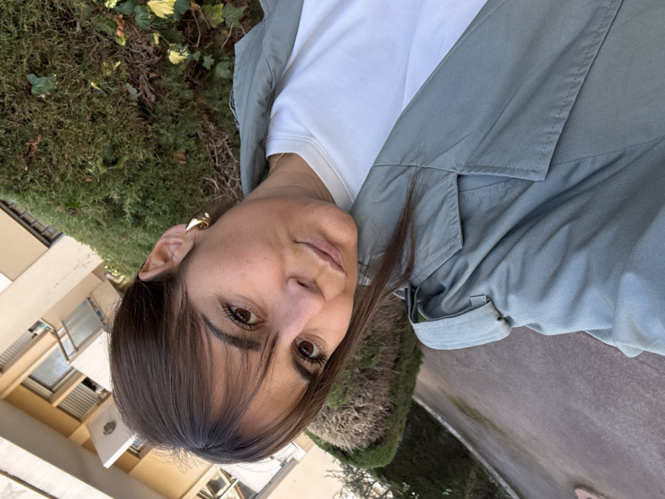 The image displays a close-up selfie of a young adult female captured in the city of Cannes, France. The woman has brown hair styled with bangs and pulled back at the sides. She has brown eyes and a neutral facial expression, looking directly into the camera. She wears a white crew-neck t-shirt and a light sage green trench coat, identifiable by its visible lapel and shoulder epaulet. A gold-toned, twisted hoop earring is visible in her right ear.
The background on the right side of the frame features dense green foliage, consistent with a well-maintained hedge, interspersed with some ivy. The left and upper background reveals a light-colored, multi-story building, likely an apartment or residential structure, featuring multiple windows, some equipped with external roller shutters, and white-railed balconies. A paved surface, possibly a driveway or pathway, occupies the lower-left portion of the scene. The lighting suggests natural daylight.