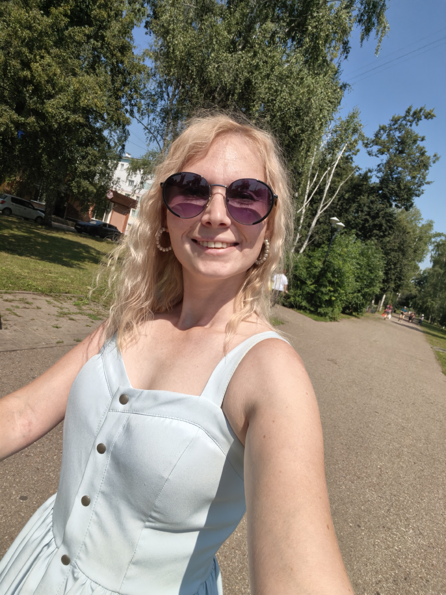 The image is a selfie taken outdoors in a park-like setting, likely on a sunny day. The subject is a blonde woman with curly hair wearing large, dark-lensed sunglasses and pearl hoop earrings. She is smiling and wearing a light blue dress with button detailing. The background shows a paved pathway, trees, grass, and some buildings in the distance, suggesting an urban or suburban environment. Several other people can be seen walking on the pathway in the background. There are parked cars on the left side of the image, and trees line the pathway. The sky is clear and blue. The location is the city of Sterlitamak, Russia.