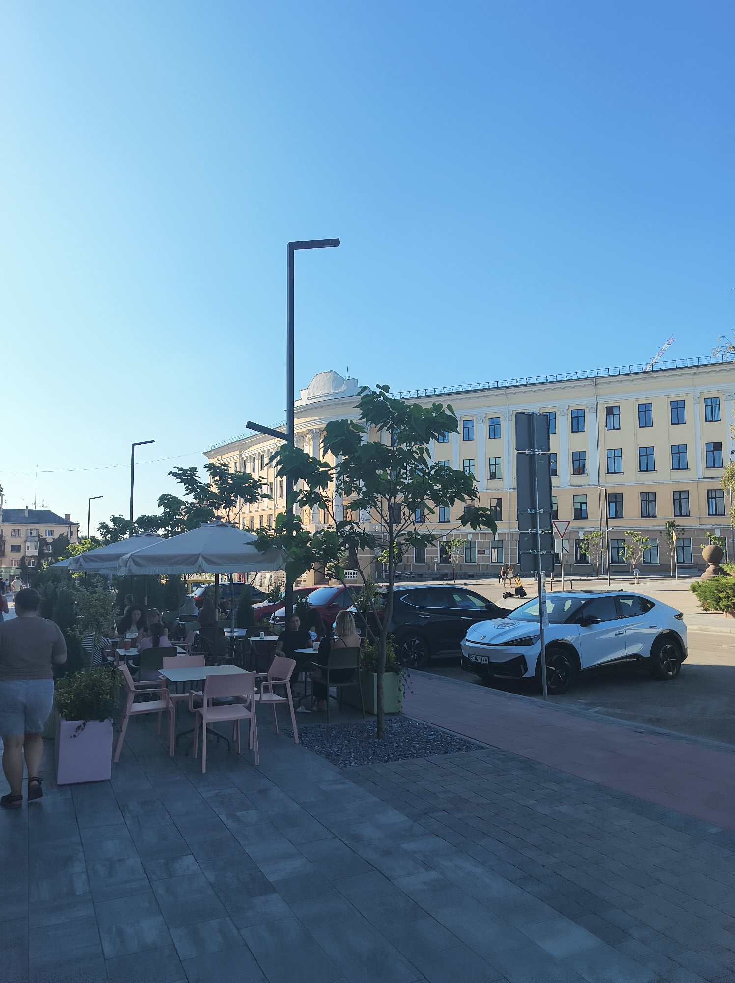 The image depicts an outdoor seating area of a cafe or restaurant in Ternopil, Ukraine, with a large, light-yellow building serving as a backdrop. The building features a repeating pattern of windows and ornate architectural details, including columns and a dome-like structure on the roof. In the foreground, several tables and chairs are set up on a paved patio, some covered by large, light-colored umbrellas. A few people are seated at the tables, engaged in conversation or dining. To the right, two cars are parked on the street: a black SUV and a white electric vehicle. A person is walking away from the camera on the left side of the image, wearing shorts and a t-shirt. Trees and shrubs are scattered throughout the scene, adding greenery. The sky is clear and blue, indicating a sunny day. The overall atmosphere suggests a public urban space, likely a popular spot for relaxation and socializing.