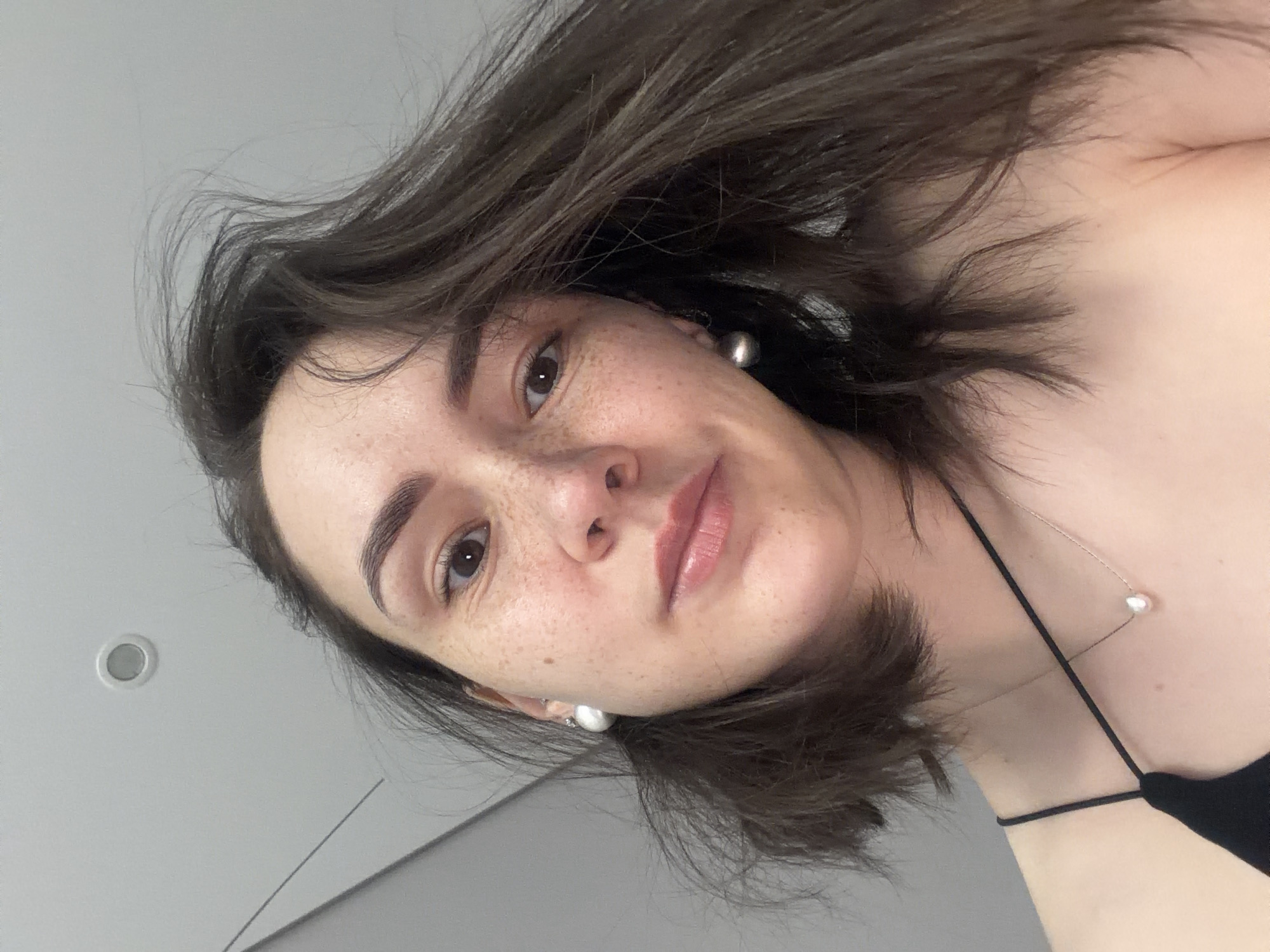 A woman with brown hair, freckles, and brown eyes is looking down at the camera. She is wearing pearl earrings and a thin-strapped black top. She also has a necklace with a pearl pendant. The background is a light gray wall with a ceiling light. The location cannot be determined from the image, and the description of Chaikovsky, Russia, is not relevant to the scene.