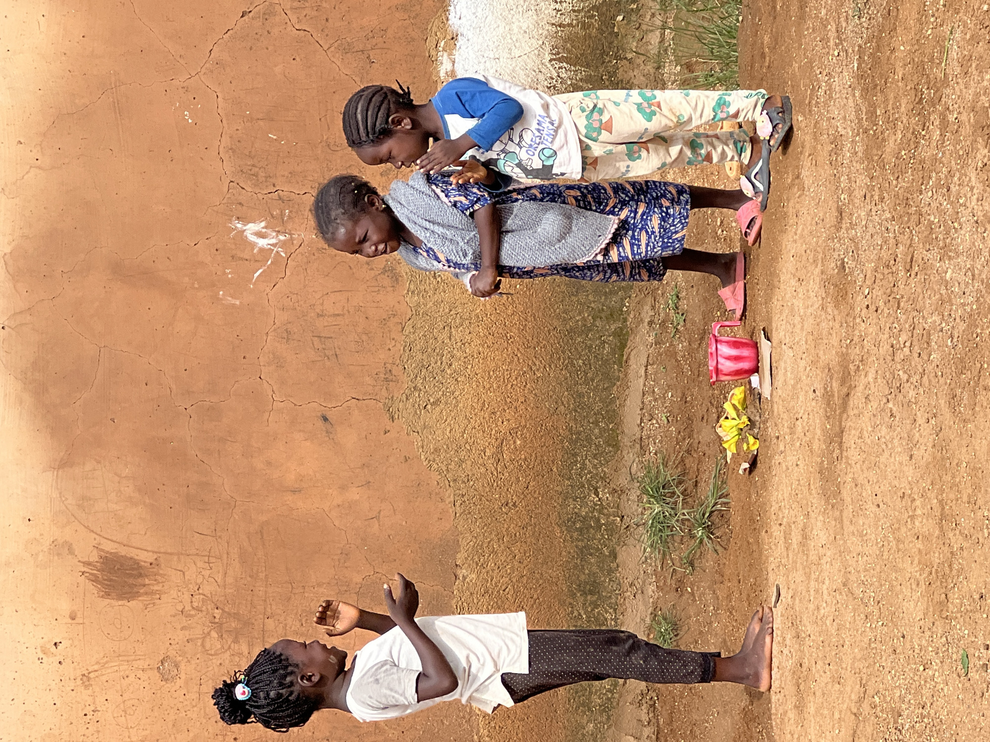 Three young Black girls are standing in front of a rough, orange-colored wall in a dry, dusty outdoor environment, likely in the city of Jos, Nigeria. The girl on the left, facing right, has her hair in braids styled in a high bun and wears a white t-shirt and dark, patterned leggings. Her hands are raised, and she appears to be speaking or gesturing. The middle girl, facing forward, wears a blue and white patterned dress with a grey knitted shawl draped over her shoulders. Her hands are clasped in front of her. The girl on the right, also facing forward, wears a blue and white long-sleeved shirt with a graphic on the front and light-colored patterned pants. Her hands are clasped in front of her, and she is looking to her left. In the foreground, on the ground between the girls, are a crumpled yellow object, a pink mug, and a few pieces of paper. The ground is predominantly brown earth with scattered dry grass and small pebbles. The wall behind them has visible cracks and some faint markings, possibly drawings or graffiti.