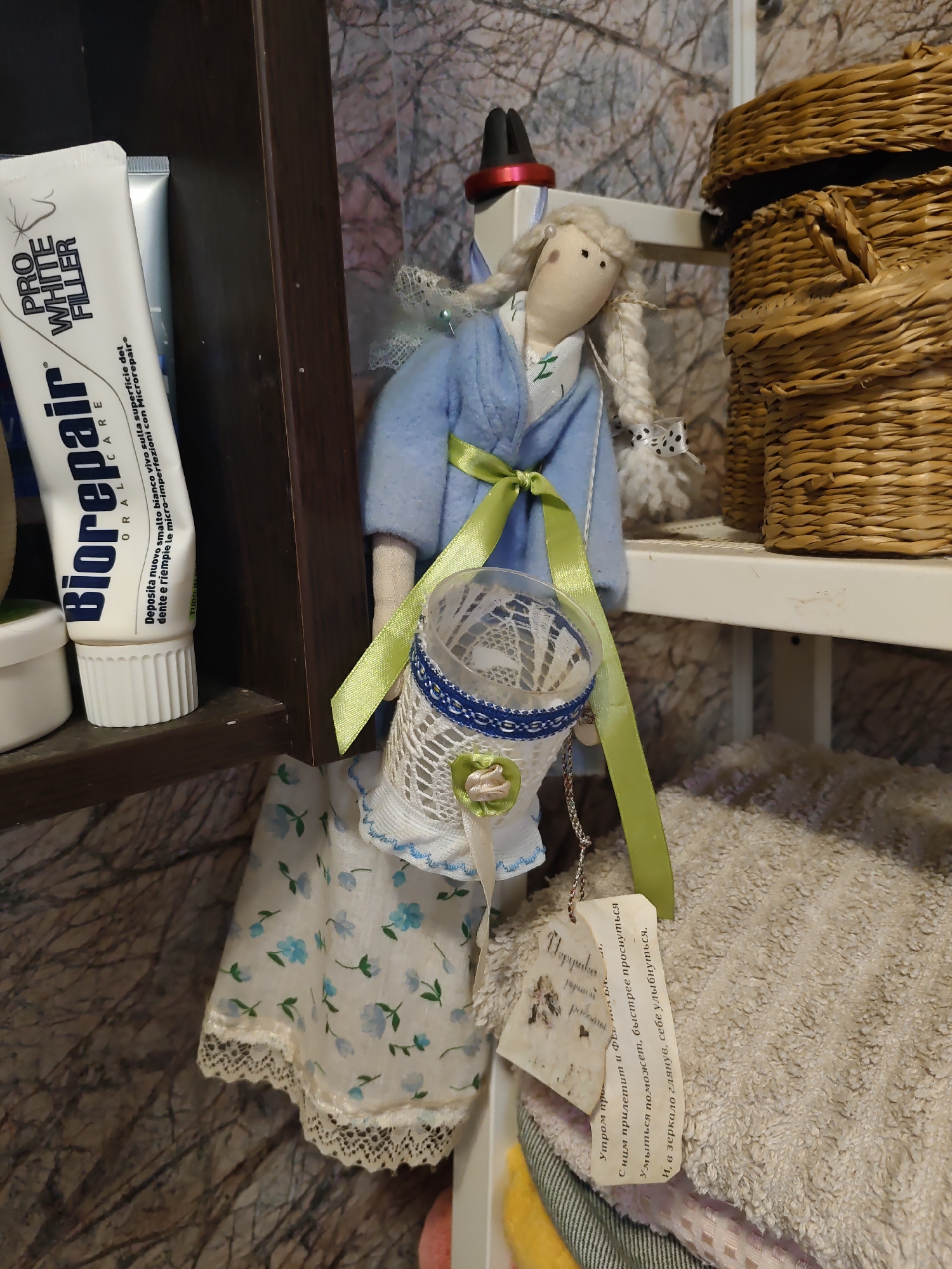 The image depicts a close-up view of a shelf in a domestic setting. On the left, a tube of "Biorepair" toothpaste is visible, positioned on a dark brown shelf. To the right of the toothpaste, a handmade doll with braided blonde hair, wearing a blue robe and a white dress with a floral pattern, is displayed. The doll is holding a decorative basket made of white lace and blue ribbon. A green ribbon is tied around the doll's waist, and a small, tag with Cyrillic script is attached to the basket. In the background, a woven basket is placed on a white shelf, and a textured wallpaper adorns the wall. The overall scene suggests a personal collection or display of handcrafted items. The image was taken in the city of Rostov-on-Don, Russia.