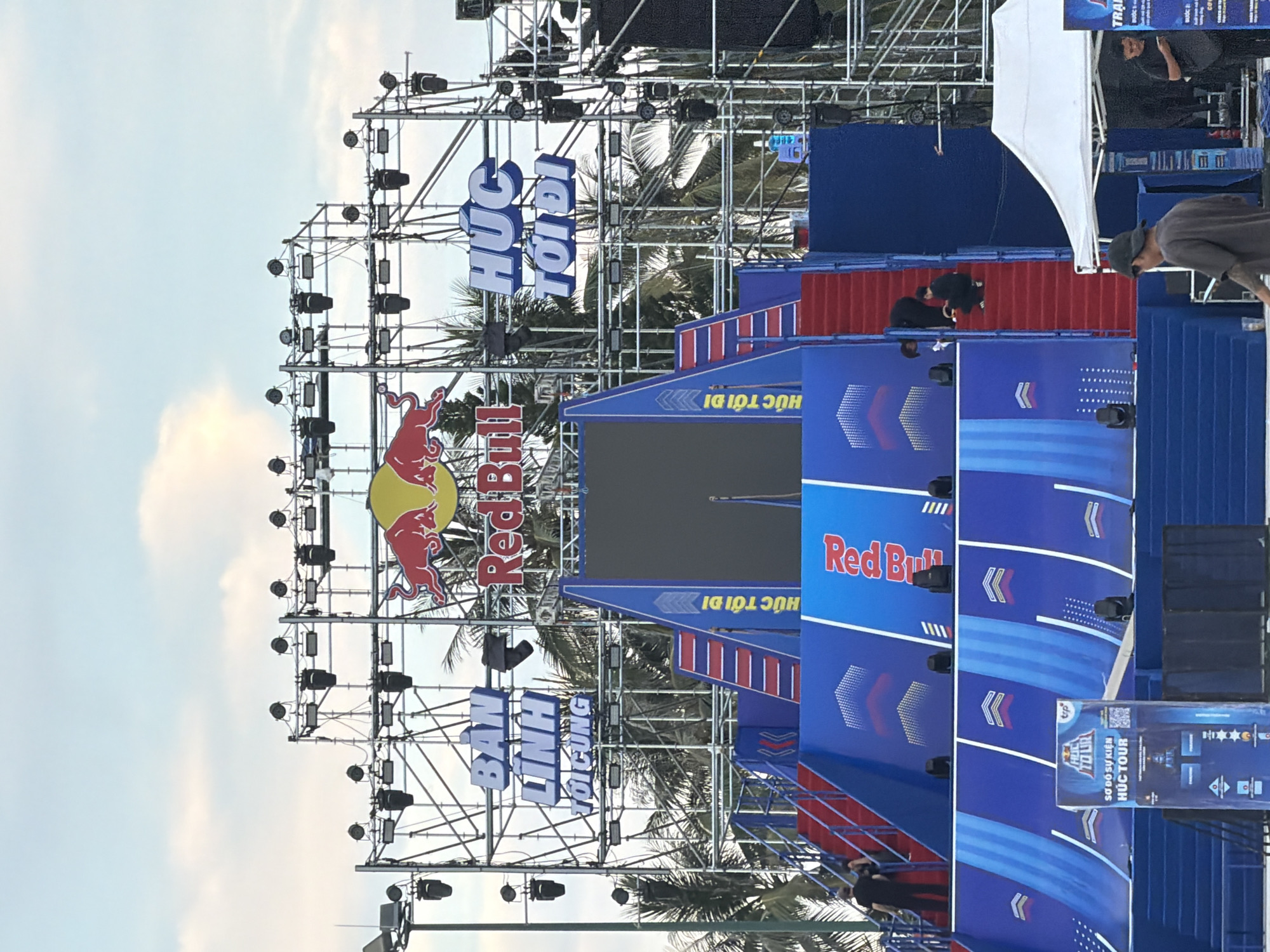 The image depicts an outdoor event setup in Nha Trang, Vietnam, likely for a Red Bull sponsored activity. The main structure is a large, multi-tiered ramp or stage with bright blue and red accents, featuring stairs and a large screen at the top. The Red Bull logo is prominently displayed on the screen and on banners. Above the stage, a complex metal scaffolding structure holds numerous stage lights. Two large signs with Vietnamese text, possibly event slogans, are mounted on the scaffolding. Palm trees are visible in the background, suggesting a coastal or tropical location. Several people are present, including individuals walking on the stairs and one person crouching near the top of the ramp. A white tent is set up to the right of the main structure, and another person is seen standing near it. The sky above is partly cloudy.