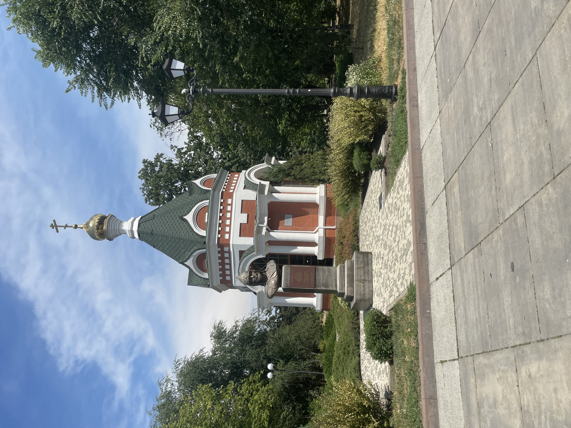 In the city of Azov, Russia, a small red and white chapel with a green roof and a golden dome stands under a blue sky with scattered clouds. In front of the chapel, a bronze bust of a man wearing a helmet and armor rests on a stone pedestal. The bust is inscribed with text. A black lamppost with two lights stands to the right of the chapel. The area is landscaped with green bushes, grass, and a paved walkway. The foreground shows a gray concrete sidewalk.