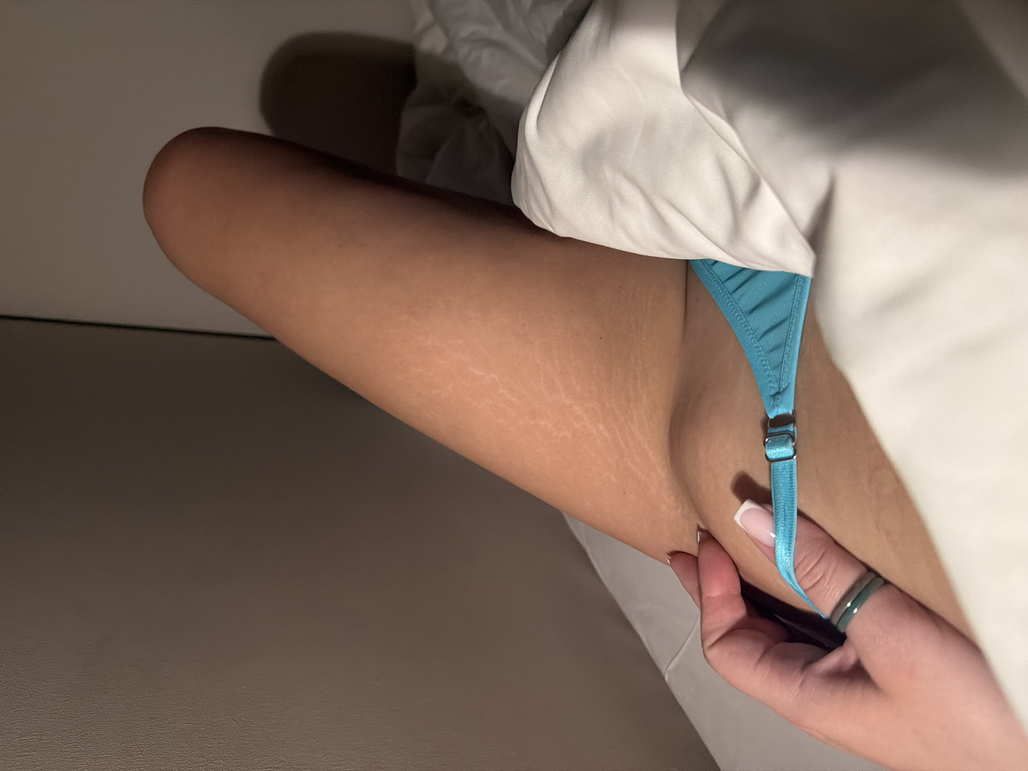 The image shows a close-up of a fair-skinned person's leg, thigh area, and part of their torso, seemingly in a bed. The leg has visible stretch marks. The person is wearing a light blue thong-style undergarment. A hand with manicured nails and wearing a ring is visible near the garment. The bedding appears to be white. The background is a muted brown surface. The location in Moscow, Russia is not determinable from the image.