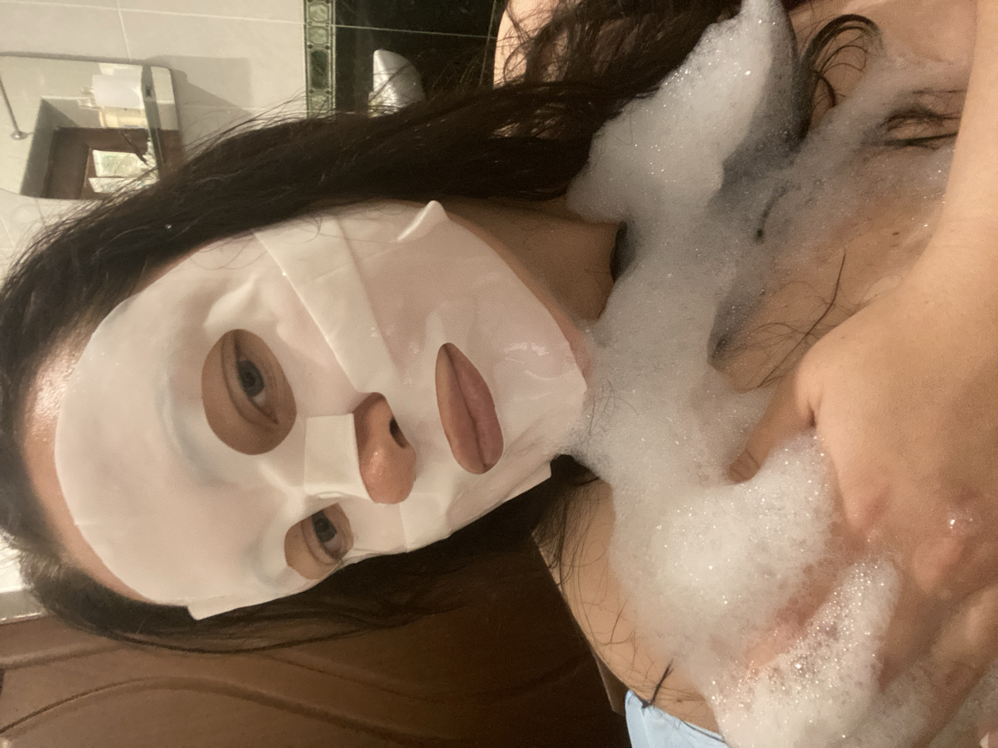A woman is in a bathroom, visible in the upper left is a reflection in a mirror and tile in the background. The woman is wearing a white sheet mask on her face and has dark hair. Foam is on her chest and partially covering her neck and shoulder. The location in the city of Sochi, Russia cannot be verified.