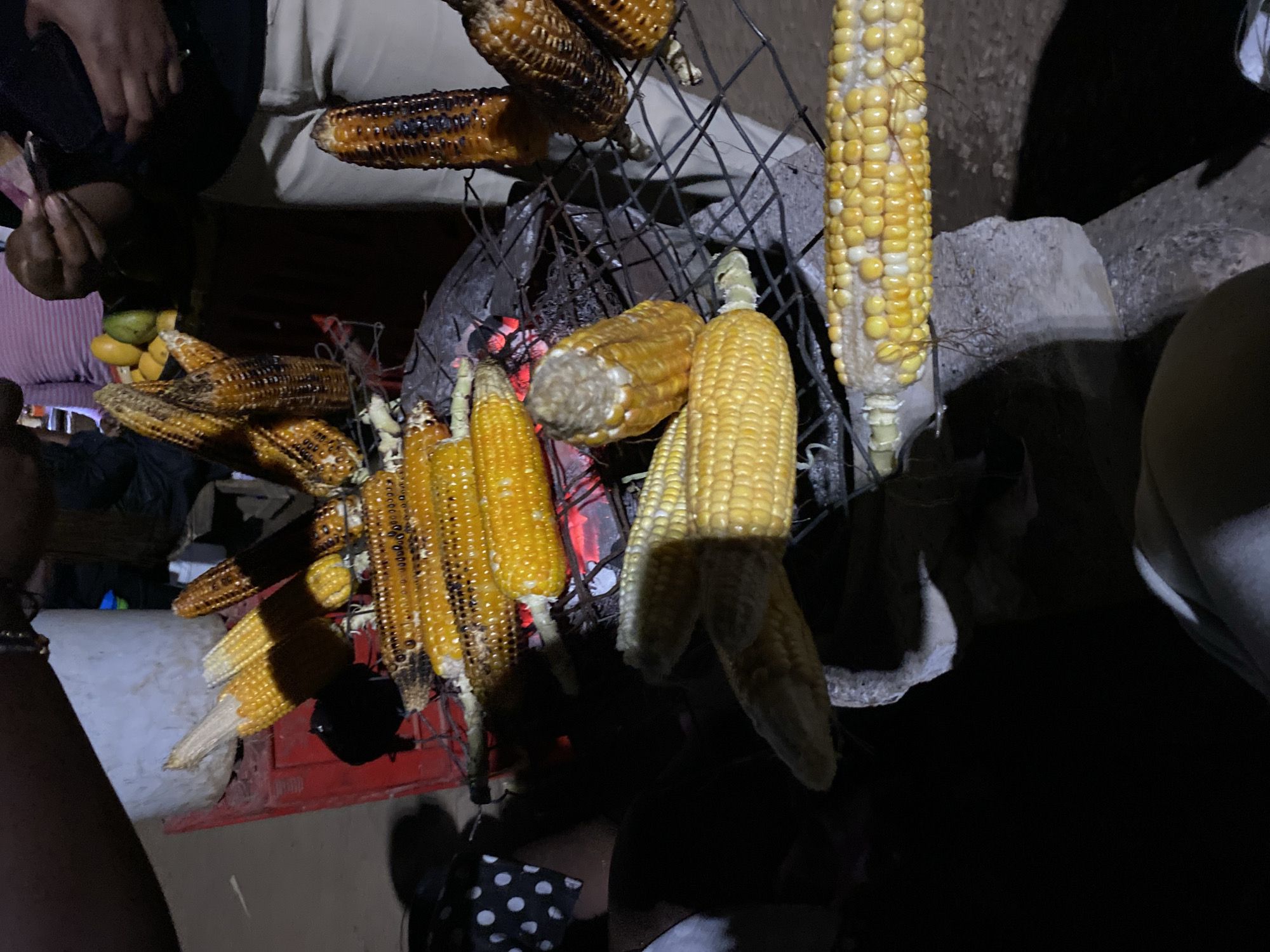 The image captures an outdoor night scene in Bukuru, Nigeria, focused on the preparation and sale of roasted corn. Multiple corn cobs are arranged on a metal grill rack directly over glowing charcoal embers, indicating they are being cooked or kept warm. Some cobs show charring and dark spots from roasting, while others appear lighter yellow and less cooked. Several raw or partially cooked corn cobs are also visible, some still attached to their husks.
Several individuals are partially visible around the grilling setup. In the upper left, a person's hands are seen holding what appears to be currency or a small item, suggesting a transaction. Another person's arm is visible on the far left, and parts of others' clothing are seen in the lower right and top center. To the left of the grill, a pile of green and yellow fruits, possibly mangoes, rests on a surface, hinting at other items being sold or consumed. The scene is illuminated by artificial light, casting bright highlights on the corn and creating deep shadows in the background. The overall darkness confirms it is nighttime. No discernible text is visible in the image.