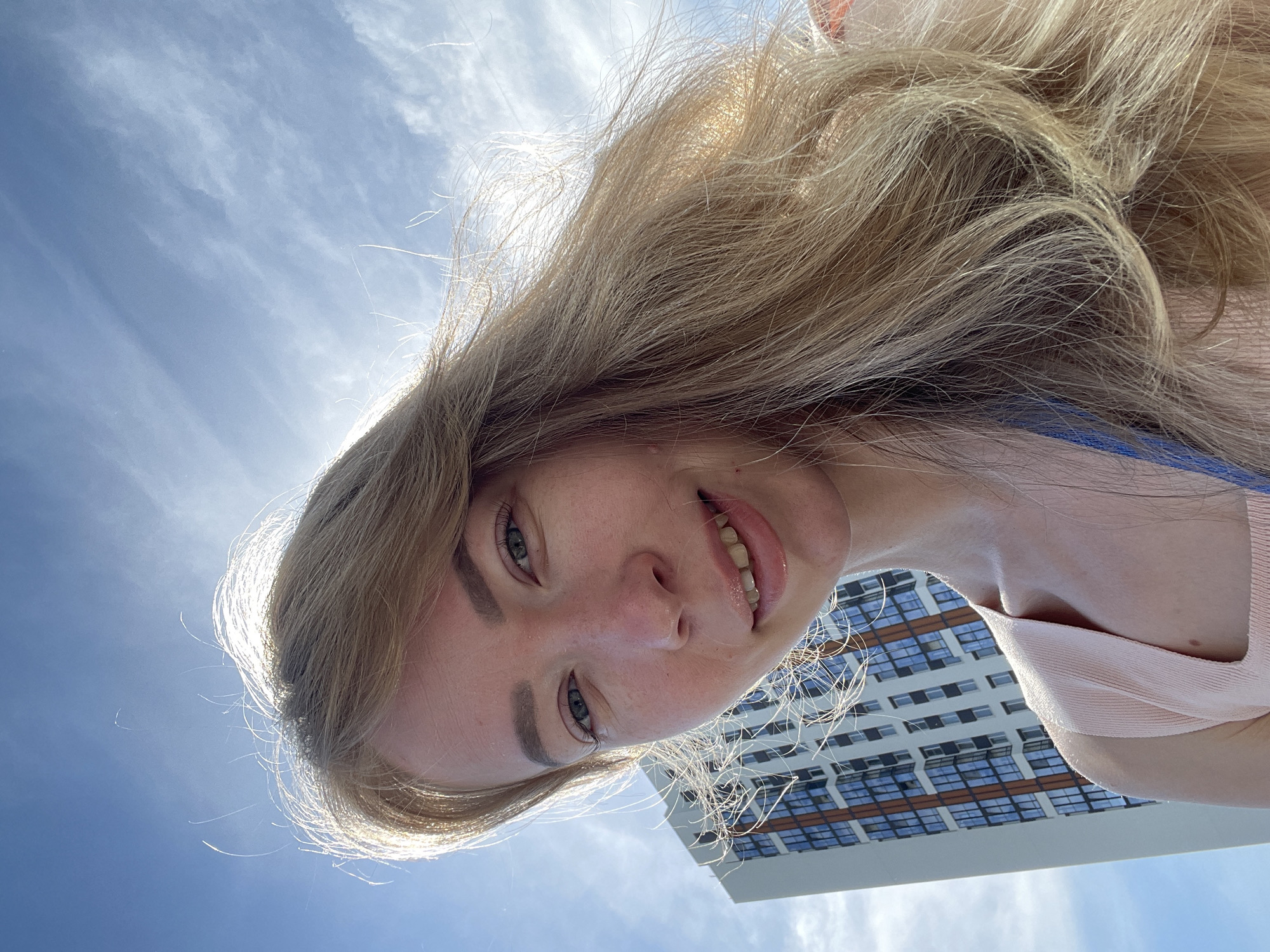 A fair-skinned woman with blonde hair and blue eyes is taking a selfie outdoors. She is wearing a light pink top. The background features a building with rows of windows and a cloudy sky. The location is likely Perm, Russia, based on the architectural style of the building. The woman is holding the camera at an upward angle. She has a slight smile.