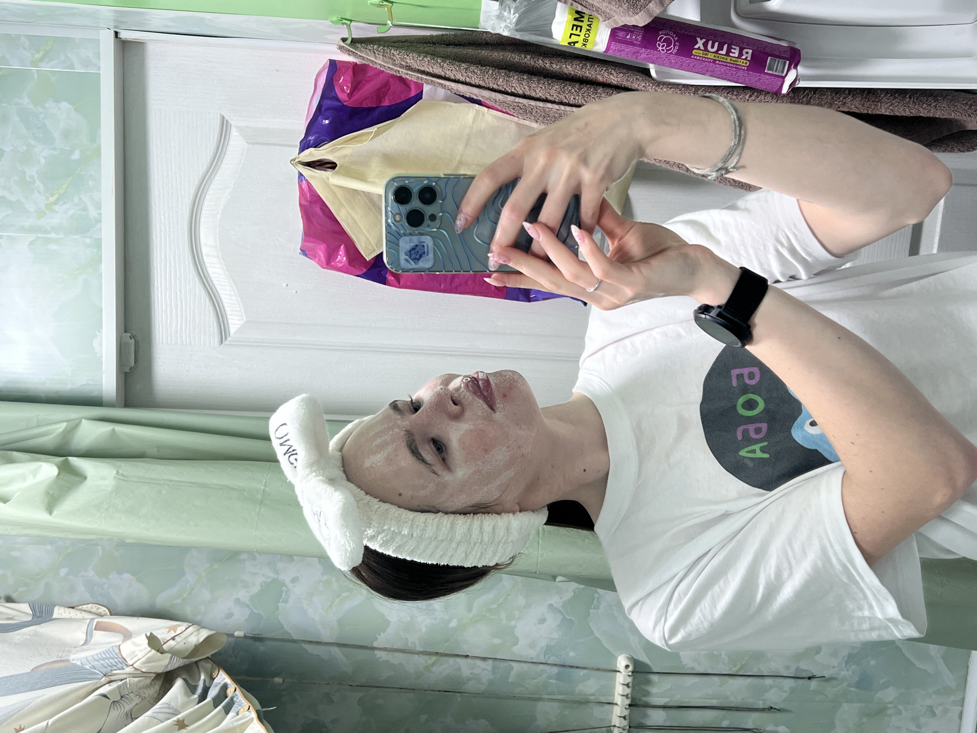 A woman in a white t-shirt with a black and green design is taking a selfie in a bathroom. She is wearing a white headband with "OMG" written on it and has a white mask on her face. She is holding a blue phone in her hands. The bathroom has green marble walls and a white door with a decorative design. There is a towel rack with various items hanging on it, including a purple bag and a brown towel. Based on the available information, it is impossible to confirm the specific location in Chaikovsky, Russia.