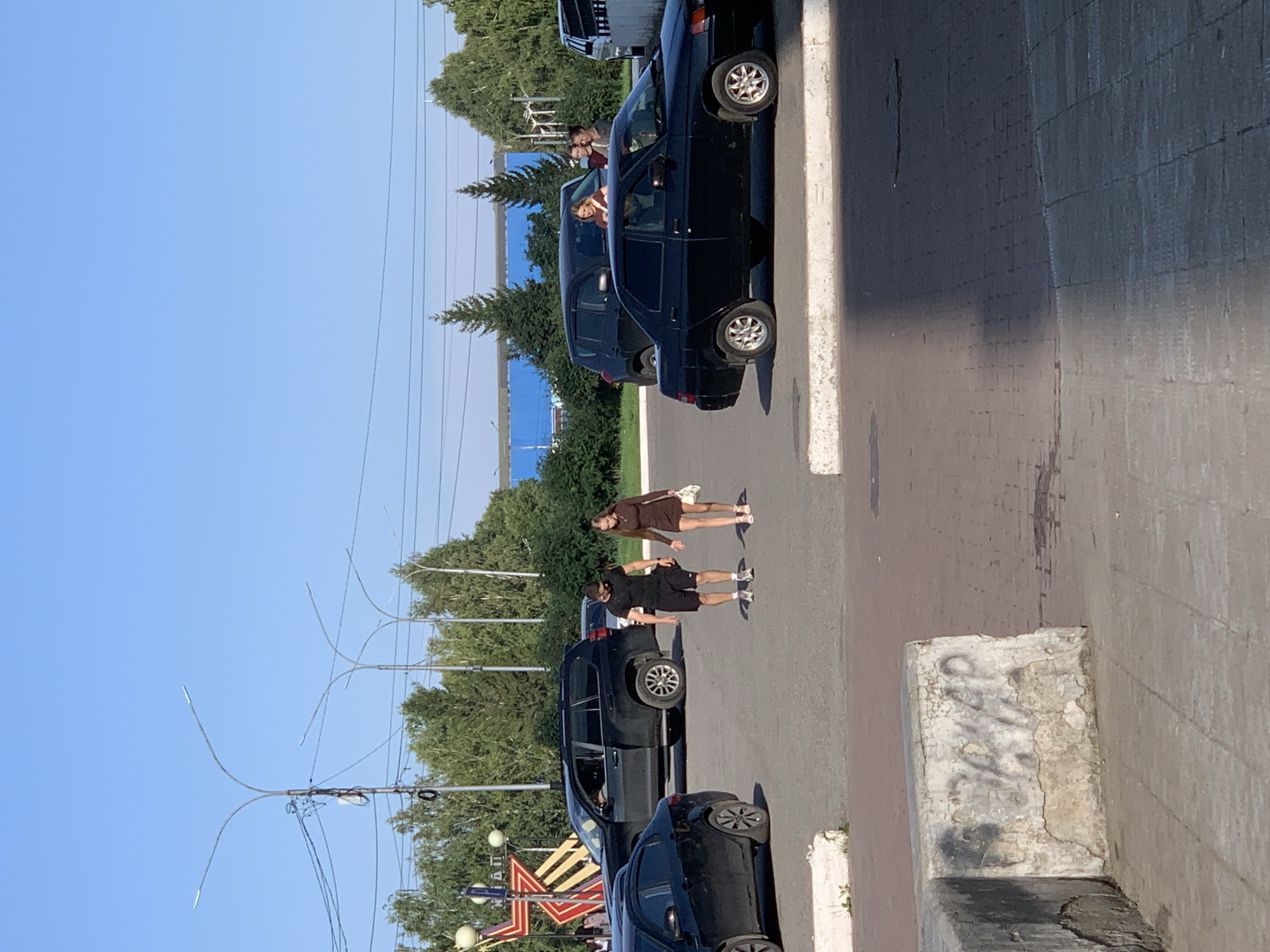 In a sunlit outdoor setting, likely a paved urban area, two individuals, a man and a woman, are walking across a parking lot or square. The man, wearing a black t-shirt and shorts, is in the process of skateboarding, as his left foot is on the board. The woman, in a brown dress, walks beside him, carrying a white purse. They are surrounded by several parked cars, including a black SUV and a black sedan, with more vehicles visible in the background. In the distance, lush green trees form a backdrop against a clear blue sky. A prominent red star-shaped monument with lights is visible to the left, suggesting a public or memorial space. The ground surface is a mix of asphalt and brick paving, with concrete curbs and barriers. The overall scene depicts a casual moment in a public urban environment.
