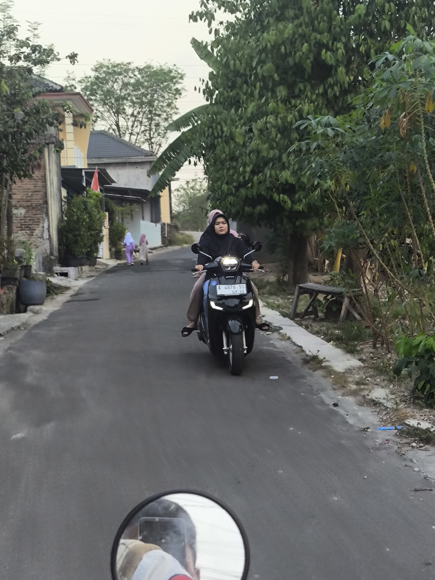 In Cilegon, Indonesia, on a paved street lined with houses and trees, a woman wearing a black hijab and a light brown shirt rides a black motorcycle. Her face is visible as she looks forward, and she is wearing blue pants and black sandals. Behind her, two women in hijabs, one light purple and one beige, walk away from the camera. The motorcycle has a license plate reading "A 4876 SS." The sky is overcast, suggesting it might be dawn or dusk. In the foreground, a motorcycle's side mirror reflects the upper body and head of the person taking the photo, who appears to be wearing a light brown vest over a white shirt. The environment is rural or suburban, with a mix of brick and painted houses, and abundant vegetation including banana trees.