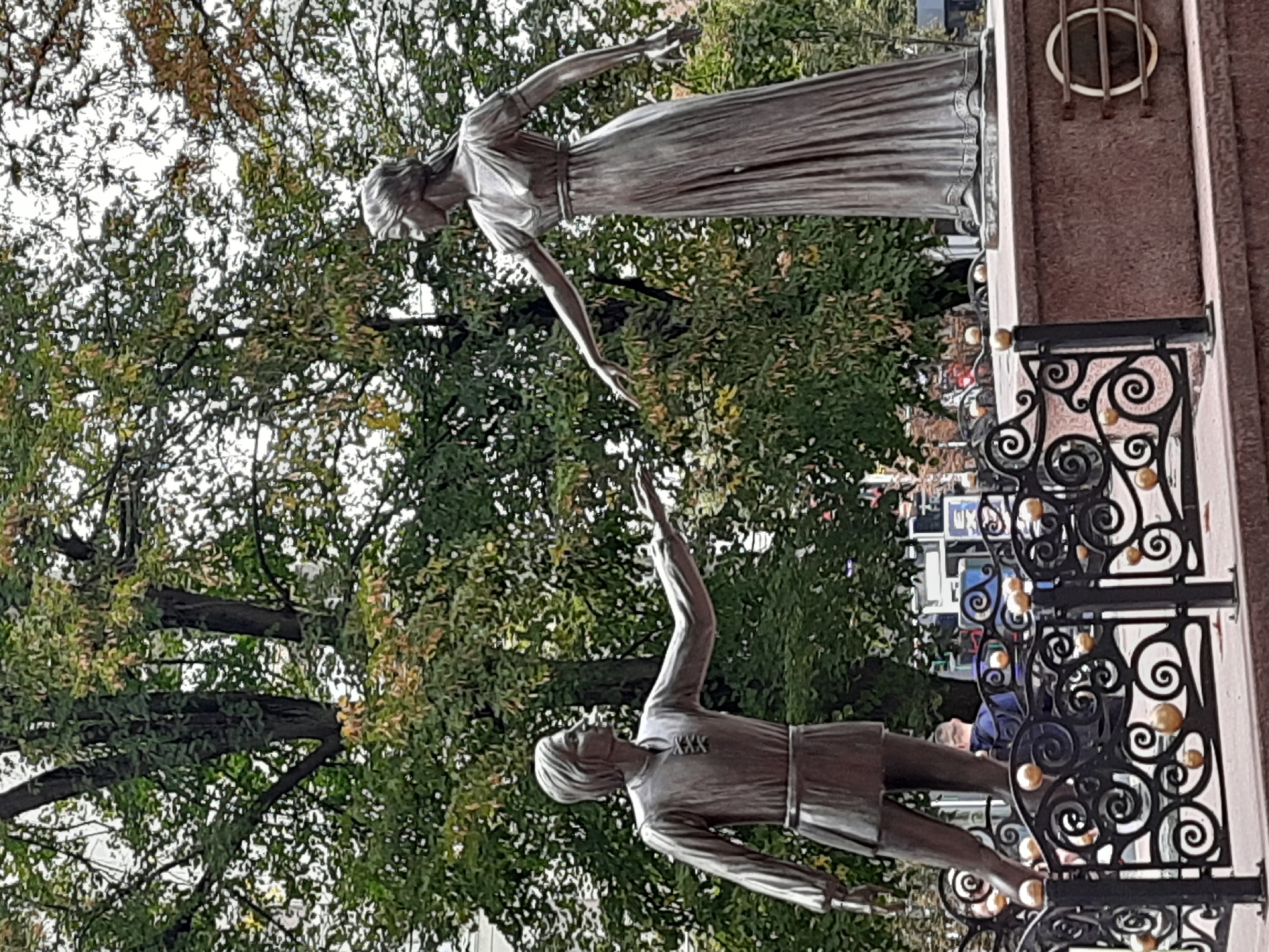 A bronze statue depicts a man and a woman reaching out to each other, symbolizing a connection or embrace. The man, to the left, wears a tunic and kilt, while the woman, to the right, is in a long, flowing dress. They stand against a backdrop of lush green trees, suggesting an outdoor setting. A decorative black metal fence with golden spheres partially encloses the statue. The statue is positioned on a raised stone pedestal. The scene appears to be in a public space, possibly a park or square. This sculpture is located in Lviv, Ukraine, specifically in front of the Lviv National Academic Opera and Ballet Theatre.