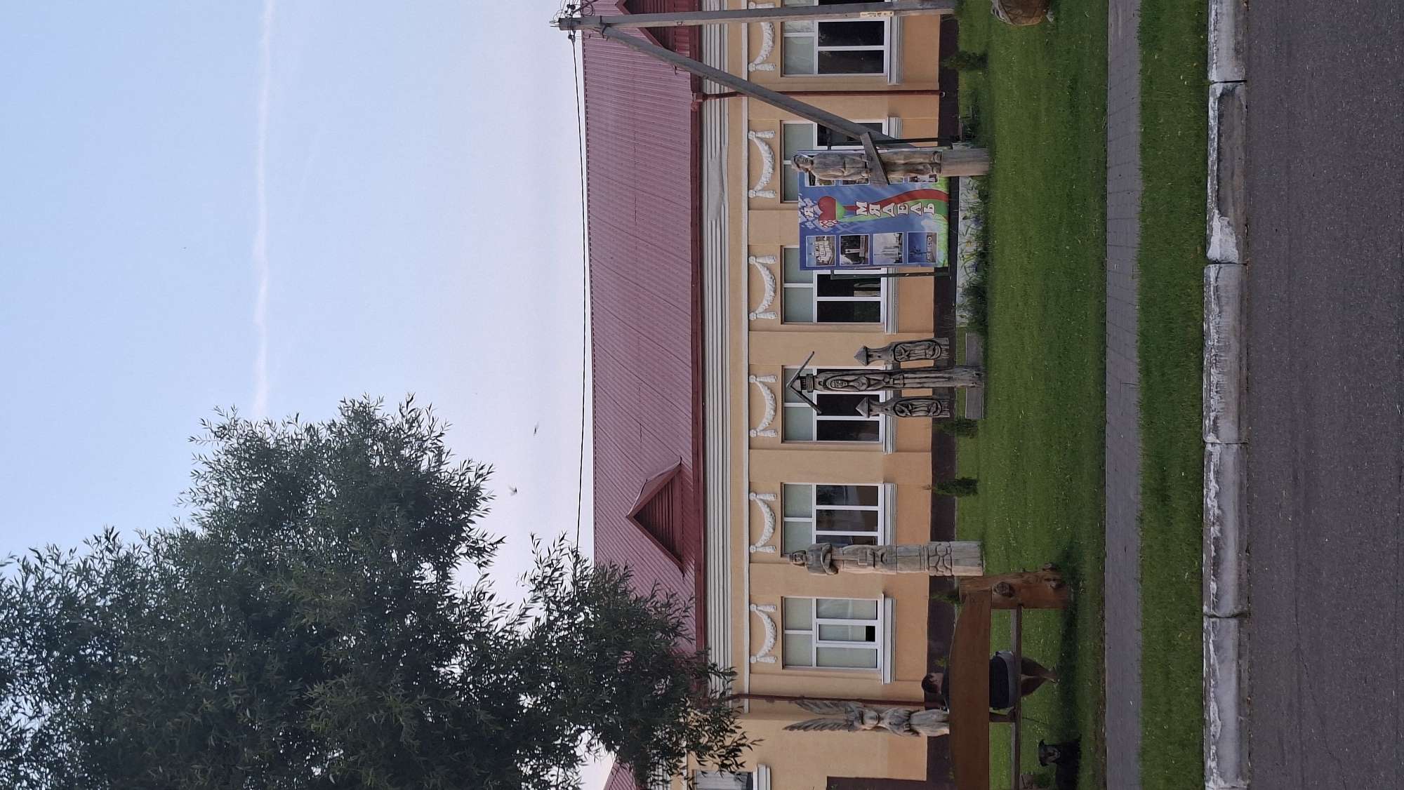 In the city of Myadzyel, Belarus, the image captures a street-level view of a building with a red-tiled roof and light yellow walls. The building features several white-framed windows and decorative molding. In front of the building, a grassy lawn is dotted with small evergreen shrubs. Several wooden sculptures are displayed on the lawn, including a tall, slender figure with carved facial features and another larger sculpture depicting multiple figures. A billboard or large poster is also visible, featuring various images and text. To the left of the sculptures, a wooden bench with a dark-colored dog sitting near it is positioned on the lawn. A large tree with dark green foliage partially obscures the left side of the image. The foreground shows a paved sidewalk and a street. The sky above is a pale blue with a faint, wispy cloud formation. A utility pole with wires stands on the right side of the scene.
