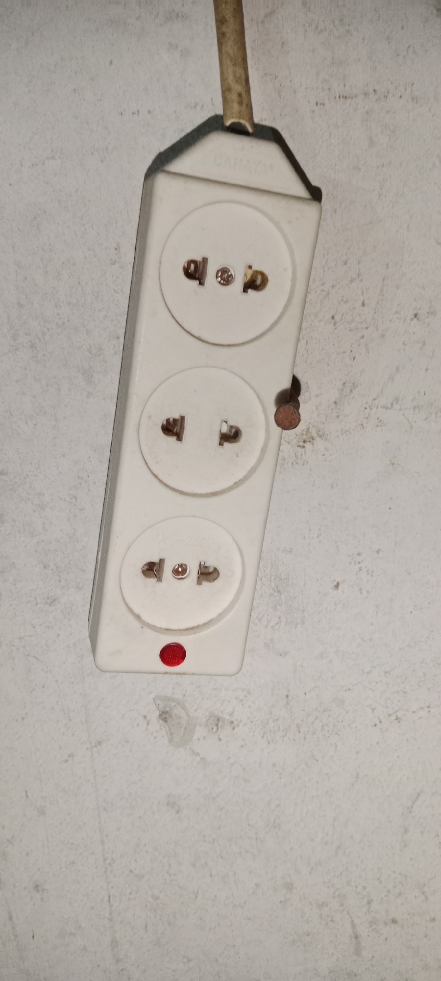 A white power strip with three sockets and a red indicator light is mounted on a textured white wall. A brown nail is partially embedded in the wall next to the power strip. A dirty, yellow electrical cord extends from the top of the power strip. The scene is indoors, likely a residential or commercial space, in Subang, Indonesia. There are no people or animals present.