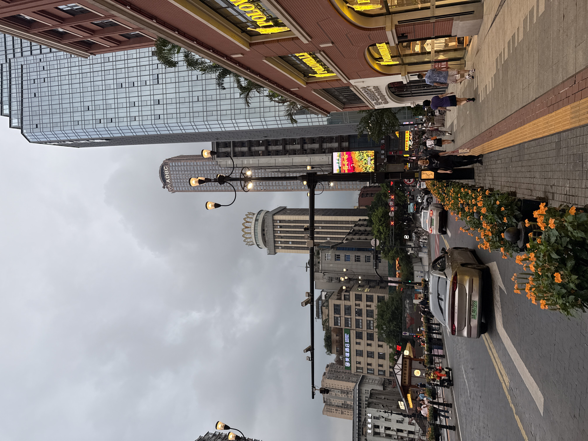 The image depicts a street scene in the Yuzhong District of China. The sky is overcast and cloudy, suggesting a dim, possibly rainy day. In the foreground, a paved street with yellow and white lane markings is visible, with a silver sedan in motion and an electric scooter with a rider. Pedestrians are walking on the sidewalk to the right of the street.
The sidewalk is lined with planters filled with orange flowers. Adjacent to the sidewalk is a multi-story building with large arched windows and signage that reads "POLOWALK" in yellow lettering. Further down the street, various other buildings are visible, including modern skyscrapers with glass facades and older, more traditional structures. One tall building features a distinctive crown-like structure at its peak. Streetlights with multiple bulbs illuminate the scene. A digital billboard displaying an image of flowers is mounted on a lamppost in the middle ground. Traffic lights and street signs are present, indicating an active urban environment. The overall impression is of a bustling city street on a cloudy day.