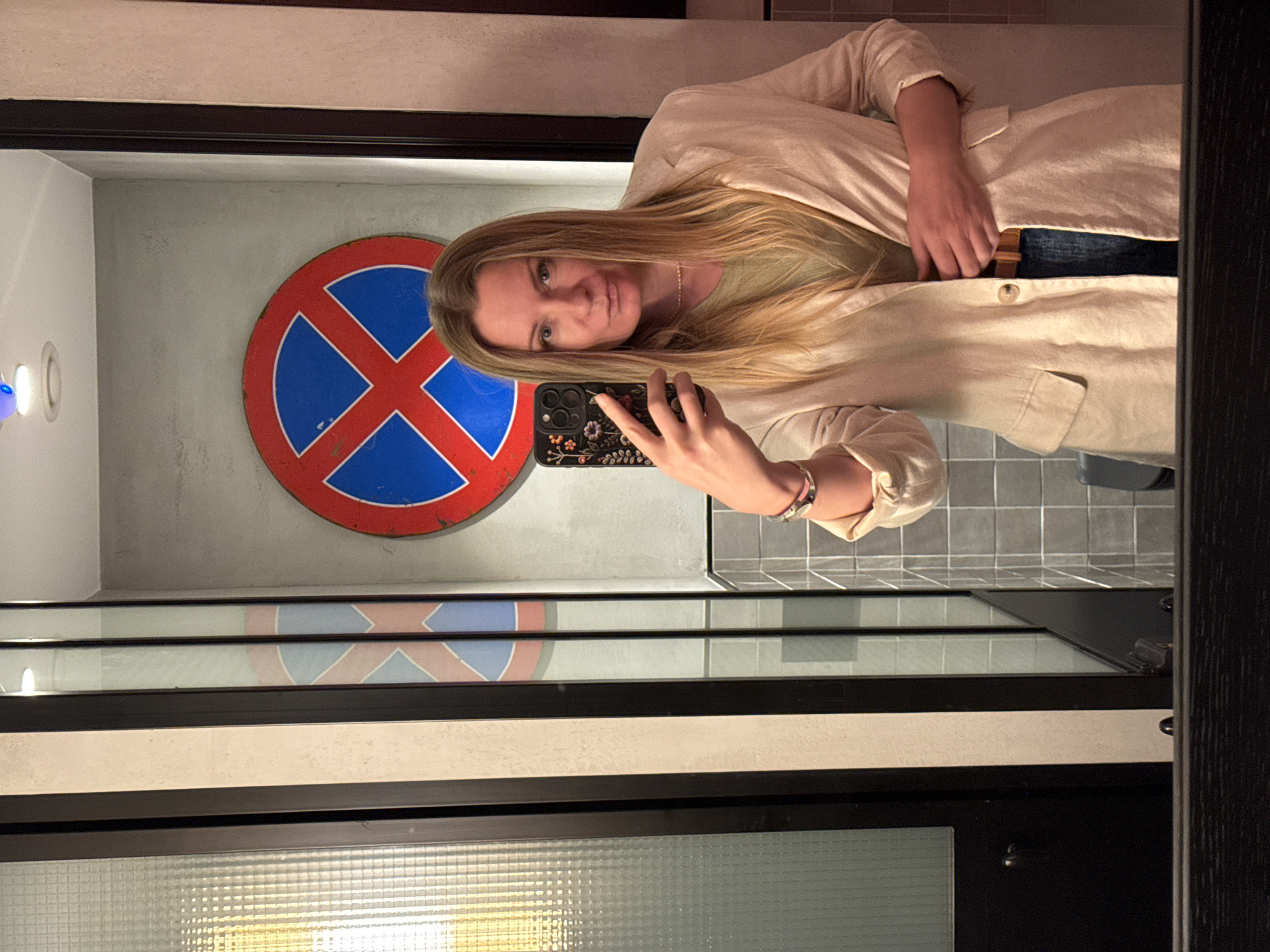 The image depicts a woman taking a mirror selfie indoors, in what appears to be Radovići, Montenegro. The woman, with long, straight blonde hair, looks directly at her phone's camera reflection with a subtle pout or smirk. She is wearing a light-colored, possibly beige or off-white, open blazer over a light green top and blue jeans. Her left hand holds a smartphone with a dark, ornate floral case, while her right hand is casually tucked near her waistband. She also wears a simple necklace and a bracelet on her right wrist.
The background visible in the mirror features a textured grey wall on which a distressed, red and blue circular sign is mounted. This sign has a red outer ring and a blue cross (X) on a white inner circle, visually resembling a stylized or vintage "no stopping/standing" traffic sign. Below this sign, the reflection shows a tiled floor and a dark, possibly wooden, frame surrounding the mirror. To the left, a light fixture emits a soft, blue-white glow. The overall setting suggests a modern interior, possibly a hotel or a contemporary living space, with an artistic or vintage decorative touch from the sign. The time of day is not discernible, but the scene is illuminated by artificial lighting.