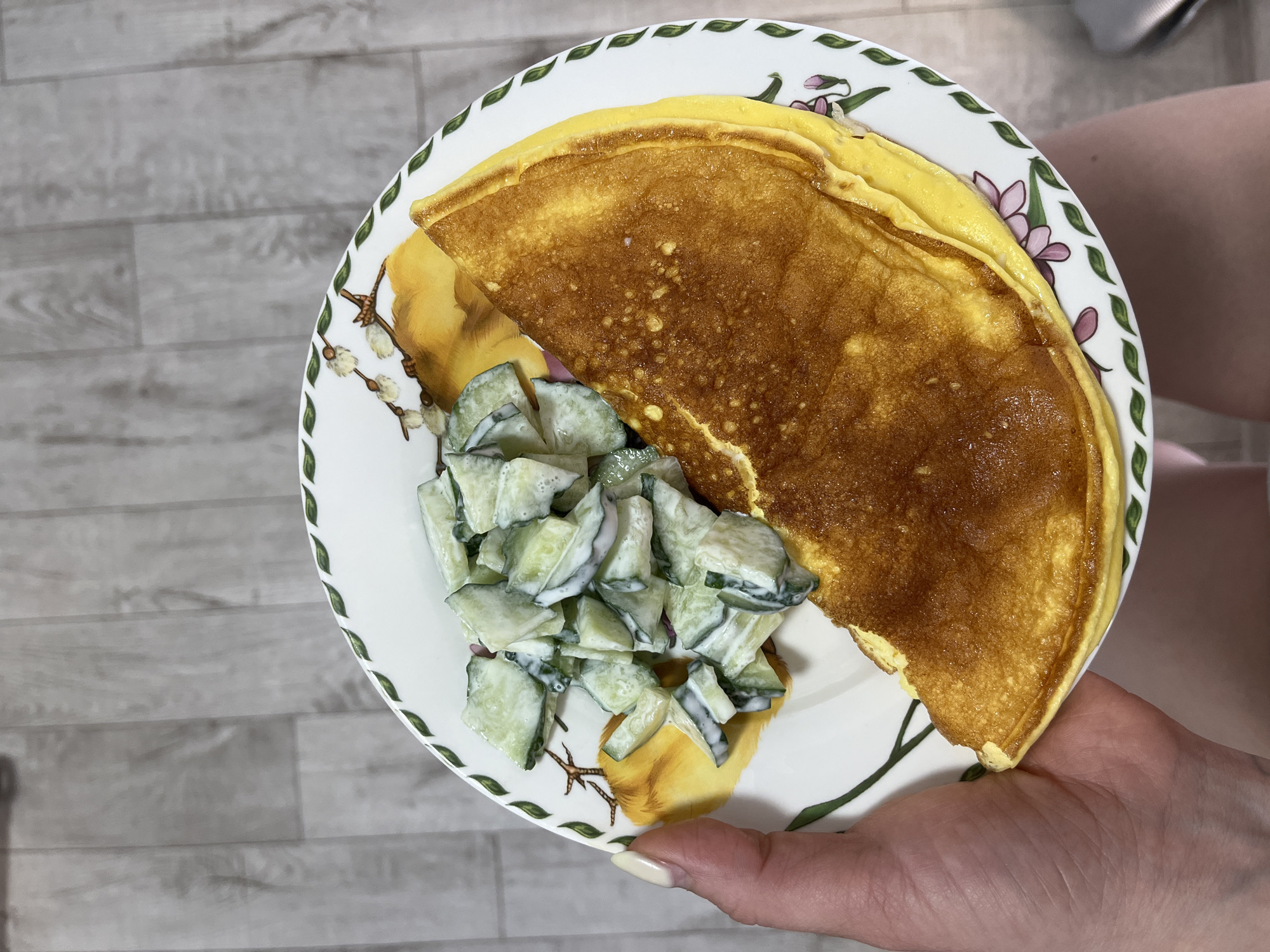A close-up, top-down shot shows a white, round plate with a floral and avian design. The plate contains a folded omelet and a side of sliced cucumbers in a creamy dressing. A person's hand, with a manicured nail, is visible holding the plate. The background is a light-colored wooden surface, possibly a floor. The scene is indoors. There is no discernible location in the city of Chaikovsky, Russia.