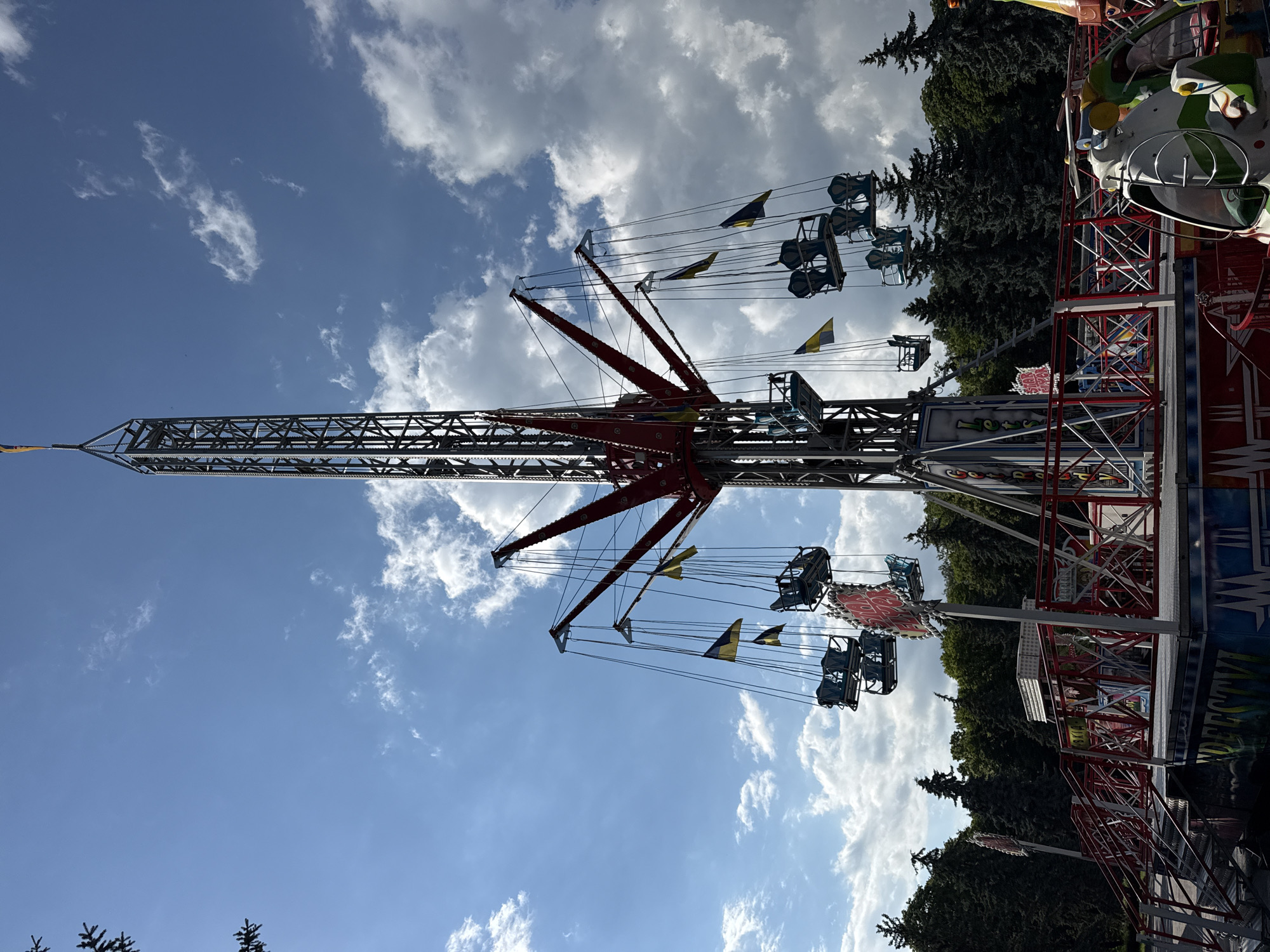 The image shows a tall amusement park ride with a metal tower and rotating arms that hold suspended chairs. Several of these chairs are visible, some occupied by passengers. Ukrainian flags are displayed on the ride. The ride is situated outdoors, with a backdrop of trees and a cloudy blue sky. The base of the ride features a colorful platform and access stairs. In the foreground, another smaller ride element, resembling a helicopter, is partially visible. The scene appears to be an amusement park or fairground in the city of Vinnytsia, Ukraine, given the presence of Ukrainian flags and the specific amusement ride.