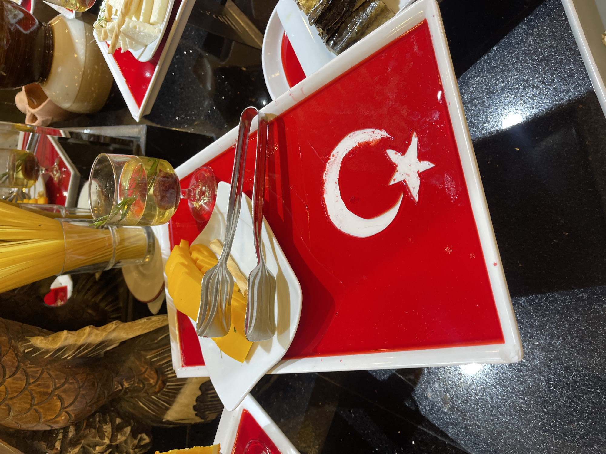 The image depicts a buffet-style food display. A white rectangular tray features a design mimicking the Turkish flag with a red background and white crescent and star. A pair of serving tongs rests on a small white plate holding pieces of yellow cheese. In the background, there are various other food items and serving dishes including a glass with a reddish tint, a jar and other trays containing food of light yellowish color. A decorative object that appears to be a carved wooden bird also lies nearby. The scene is set on a dark, speckled countertop. The location is Unknown, Unknown.