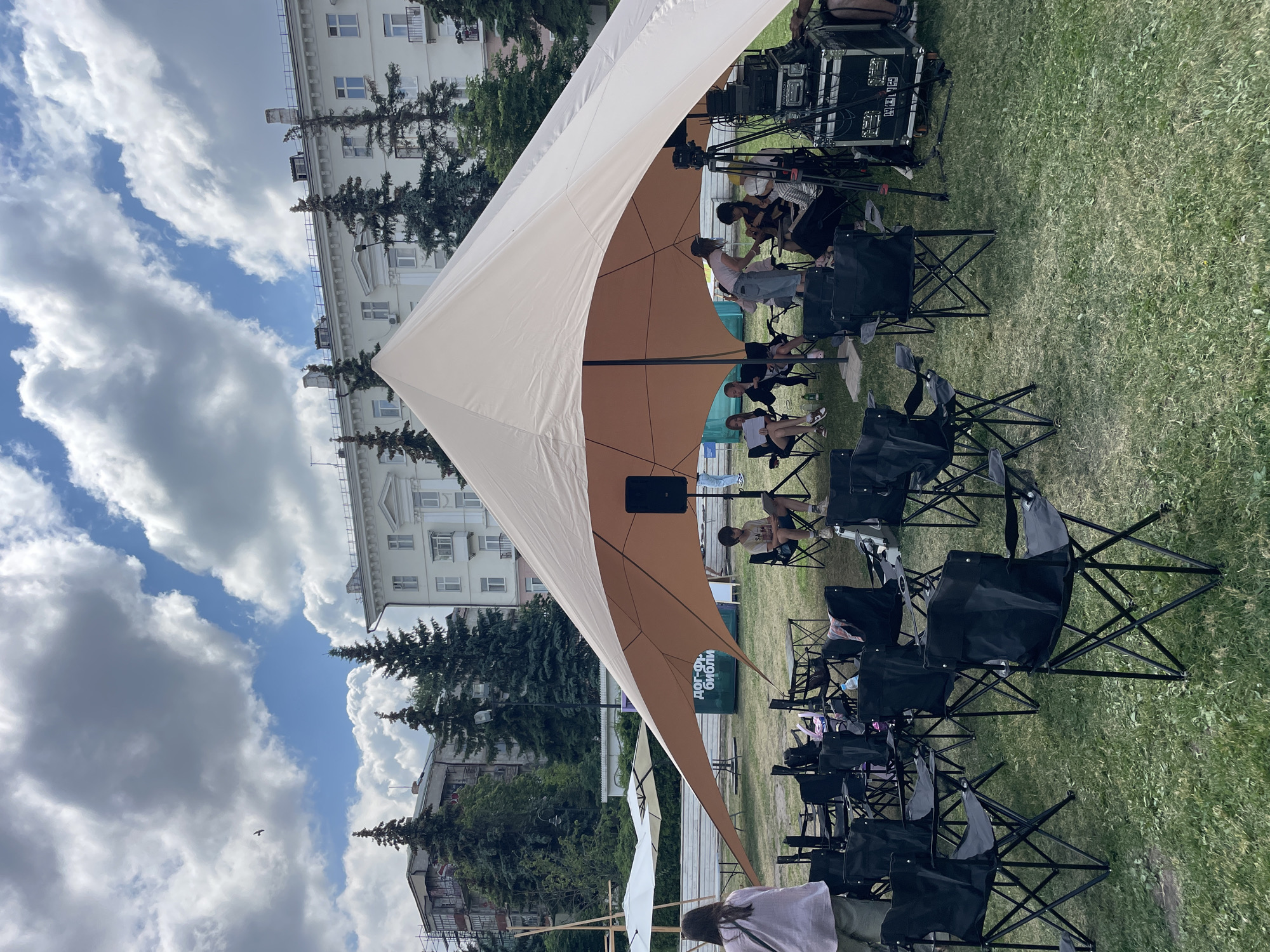 The image depicts an outdoor event on a cloudy day, possibly in the city of Kazan, Russia, given the Cyrillic text on a sign. A large, light-colored tent, likely a canopy, covers a gathering area. Numerous black folding chairs are arranged on the grass in front of the tent, some occupied by people. To the right of the tent, equipment, including a speaker and what appears to be a sound mixing console on a road case, is set up. Several trees are visible behind the tent and in the background, along with buildings. In the foreground, a woman in a pink shirt stands facing away from the camera, observing the scene. A bird is visible in the sky.