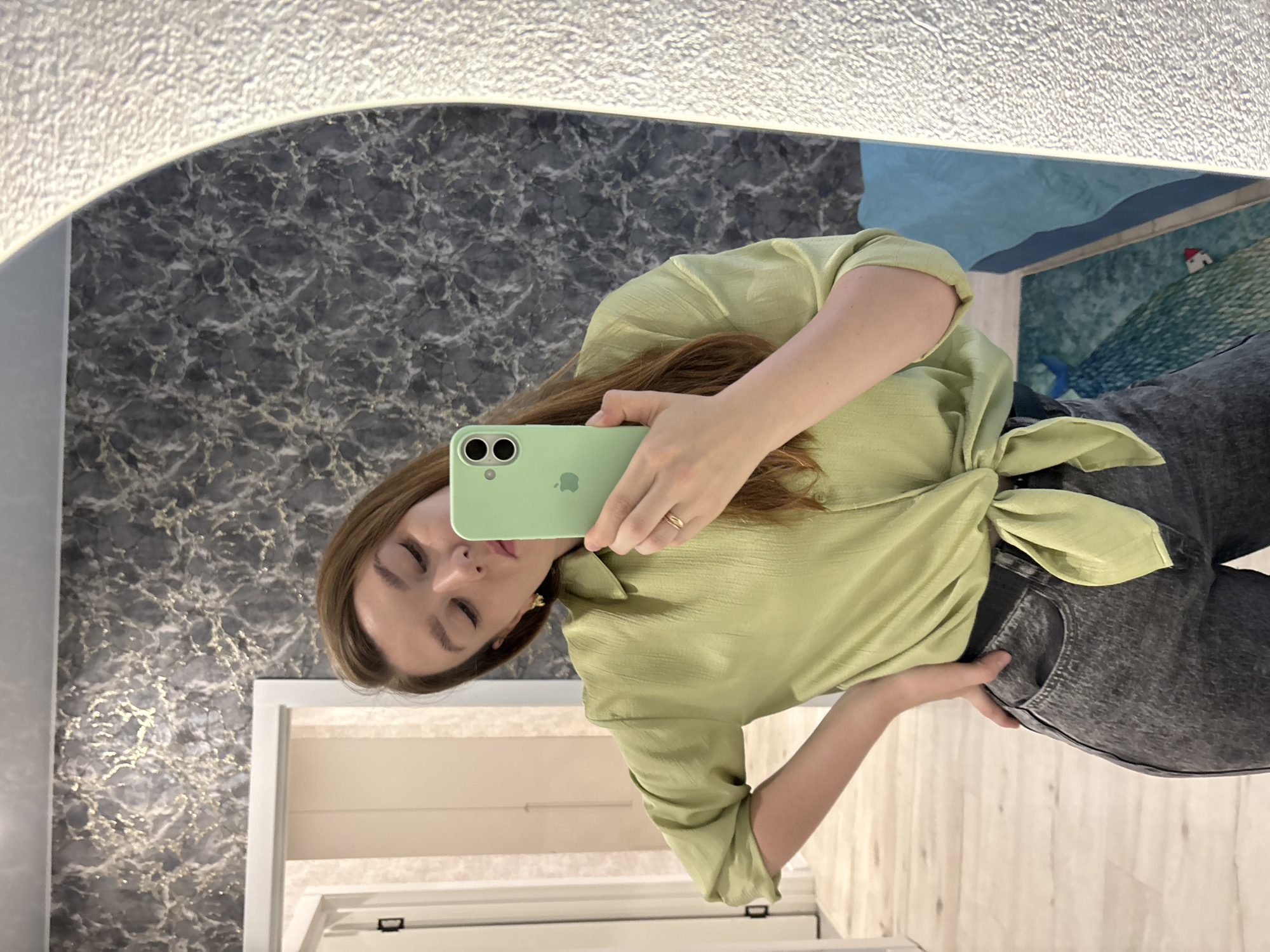 A woman is taking a mirror selfie. She is wearing a light green blouse tied at the waist and dark gray jeans. She is holding a light green iPhone. The reflection shows a bathroom with gray marble-patterned wallpaper, a mirror with a lighted frame, a painting of a sea landscape, a white door, and a light wood floor. This photograph was likely taken in a private residence in Izhevsk, Russia.