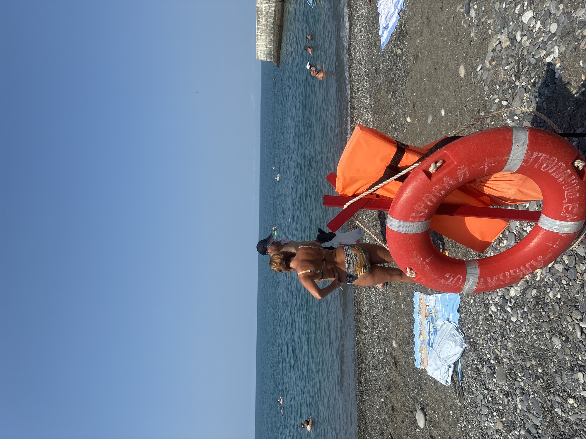 The image shows a sunny day on a pebble beach in Sochi, Russia, with the calm, blue sea in the background. A red life preserver with white reflective stripes is prominently displayed in the foreground, attached to a red pole, alongside an orange life vest. Two people, a woman in a patterned bikini and a man wearing a black cap, stand near the water's edge, facing away from the camera. Several other individuals are visible in the sea, swimming and relaxing in the water. A concrete pier or breakwater juts out into the sea on the right side of the frame. The beach is covered in small, grey and dark pebbles. A rolled-up towel with a beach scene printed on it is lying on the pebbles to the left of the life preserver. The sky is clear and bright blue.
