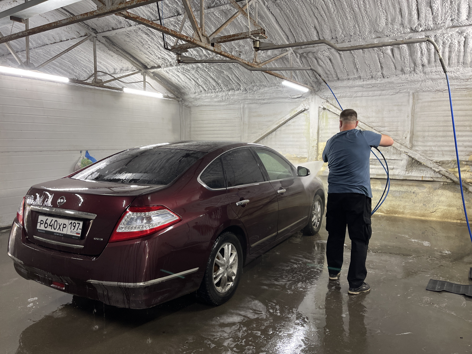 Inside what appears to be a car wash bay, a man is seen from behind, actively washing a dark red (maroon) Nissan Teana sedan with a high-pressure hose. The interior features a practical, industrial aesthetic with white, corrugated walls and a heavily insulated ceiling covered in a textured, white spray foam. Exposed metal beams, some with visible rust, form part of the ceiling structure, from which fluorescent lights and the blue hose system are suspended.
The man, dressed in a blue short-sleeved t-shirt, dark cargo pants, and Fila sneakers, holds the hose, directing a spray of water towards the car. The car is visibly wet, with water streaks and suds on its surface and pooling on the concrete floor, which reflects the artificial lighting. On the car's rear, a Russian license plate is clearly visible, displaying "P640XP 197 RUS", with "197" denoting the Moscow region. Other visible text on the car includes "TEANA" and "NISSAN" badges on the trunk, and a "V6" emblem, indicating the engine type. Small, colorful plastic bags hang on a hook on the wall to the left of the car. The overall scene suggests an indoor, utilitarian environment focused on vehicle maintenance, likely during the day or evening, as natural light is not apparent.
