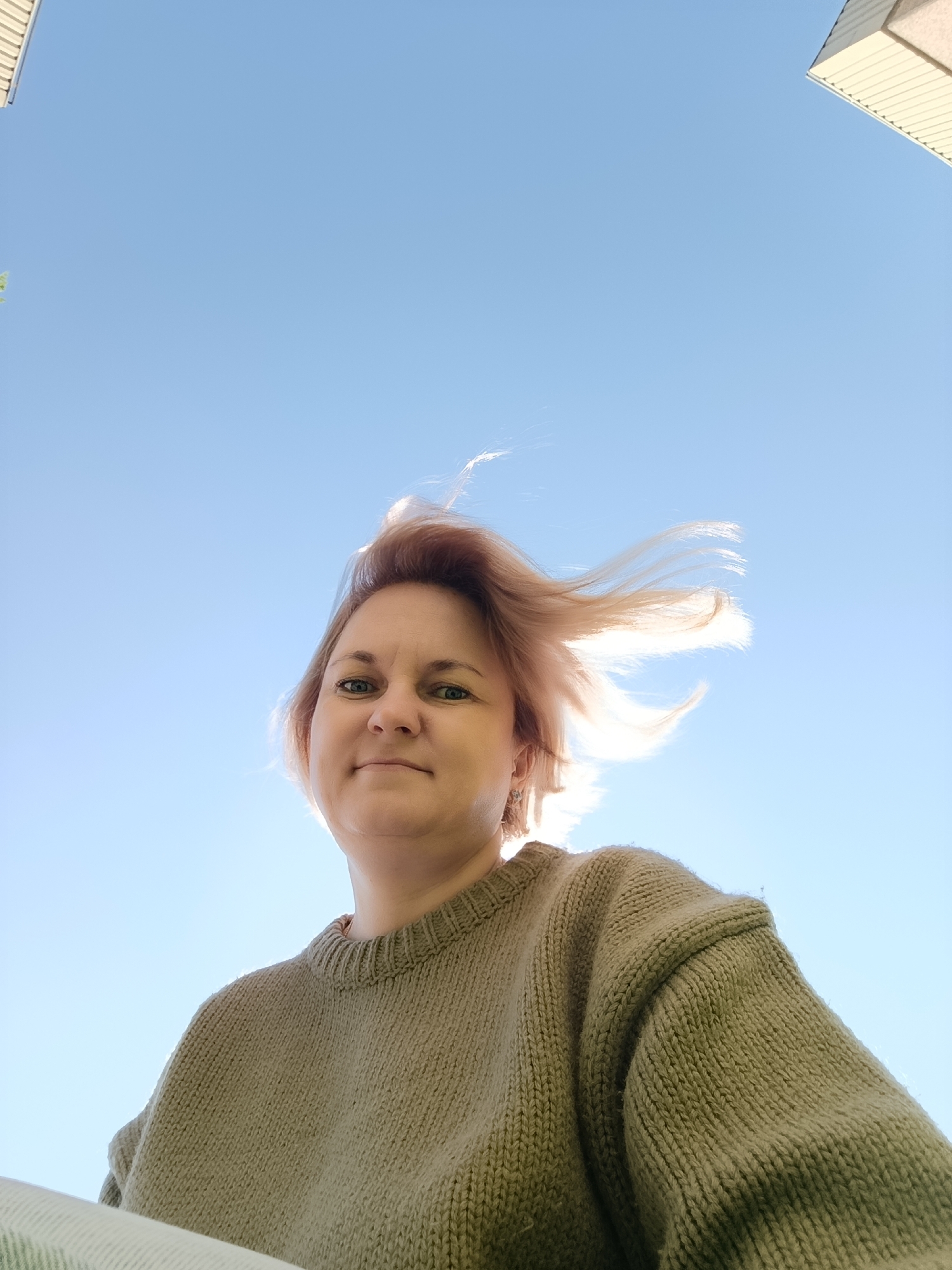 A low-angle outdoor portrait features a woman looking directly towards the camera against a clear, bright blue sky. She has short, light-colored hair, possibly pinkish-blonde, which appears wind-blown and is strongly backlit by the sun, creating a radiant halo effect around her head. Her expression is calm and pleasant, with a subtle smile. She is wearing a chunky, light brown or olive green knitted sweater with a ribbed crew neck.
The dominant background is the vibrant, cloudless blue sky. In the upper right corner, and a small sliver in the upper left, parts of a light-colored building structure, possibly a roofline or overhang, are visible. The strong backlighting and wind-swept hair suggest a sunny and breezy daytime. No text is visible in the image. The photograph was taken in an outdoor setting, possibly in Марковский, Russia.