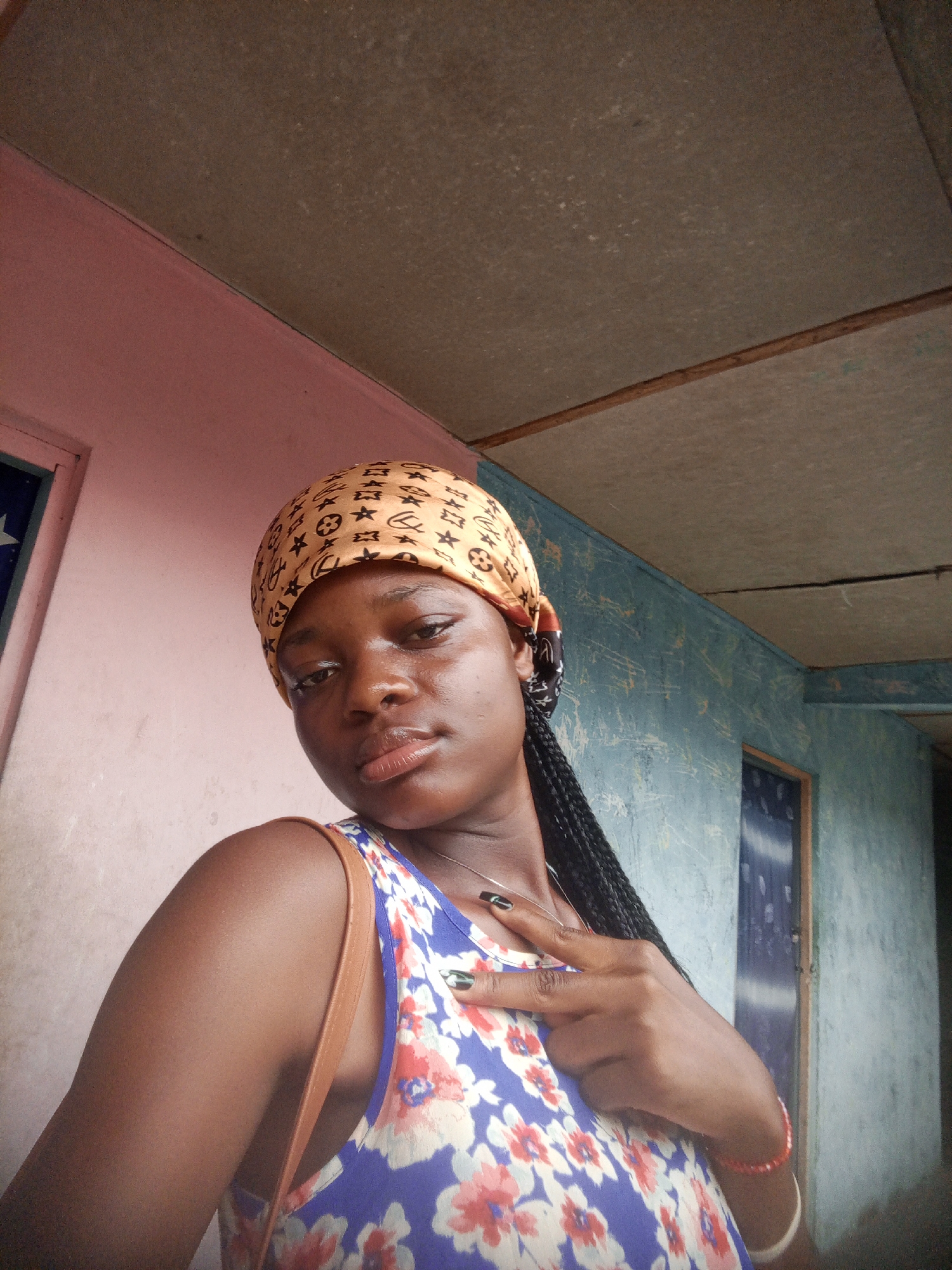 A young, dark-skinned woman is the subject of the photograph. She is wearing a headscarf patterned with a tan color. Her hair is styled in long braids that fall over her shoulder. She is wearing a sleeveless floral top with a blue background and red and white flower patterns. A brown bag strap is visible on her shoulder. She is wearing a necklace and bracelets on one wrist. Her fingernails are painted black with a silver design. She is gesturing with her hand. The background includes a building with walls painted in pale pink and blue shades. A door is visible in each of the colored wall segments. The image was captured inside a building somewhere in the city of Mbribit, Nigeria.