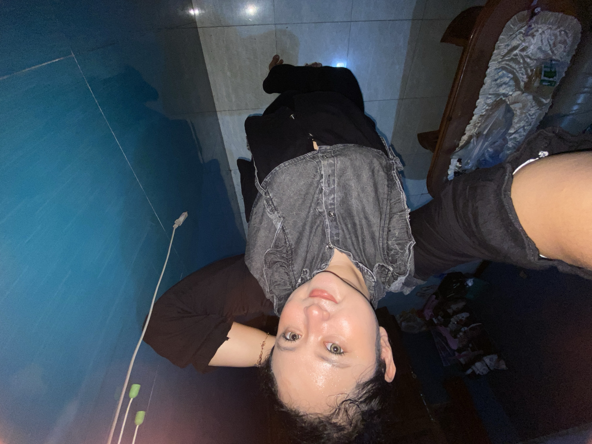 The image shows a young Indonesian woman taking a selfie. She is wearing a dark grey denim vest over a black shirt and black pants. Her hair is styled in a bun, and she has light makeup on. She is posing with her hand behind her head. In the background, there are some shelves with items on them and a blue wall. A wooden table with a plastic-wrapped object is visible in the lower left corner. The scene appears to be indoors. The image is not specific enough to determine the exact location in Gelam, Indonesia.