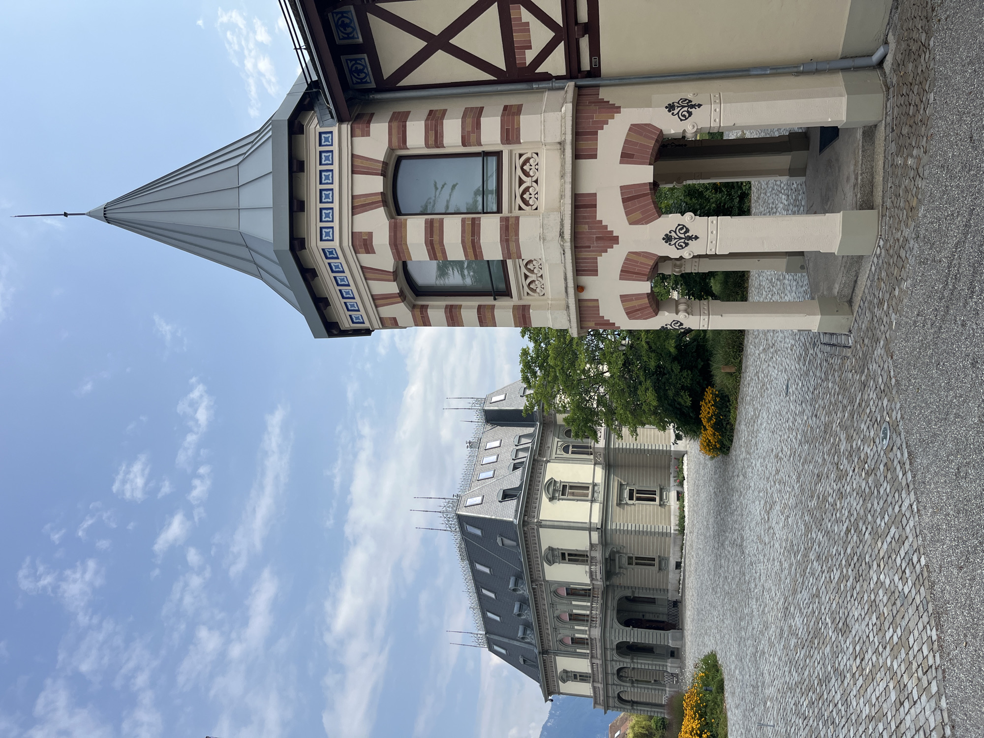 The image shows a stately mansion in La Tour-de-Peilz, Switzerland, with a cobbled driveway leading up to it. The mansion is a grand, pale yellow building with a dark grey, mansard-style roof featuring multiple dormer windows and decorative finials. To the right of the mansion, a smaller, octagonal tower with a conical roof is visible. This tower has red and white striped brickwork around its base and a band of blue and white patterned tiles near the top. The tower has arched openings at its base, supported by columns. The grounds are landscaped with green trees and patches of yellow flowers. The sky is a pale blue with scattered white clouds. The scene is calm and appears to be a sunny day.