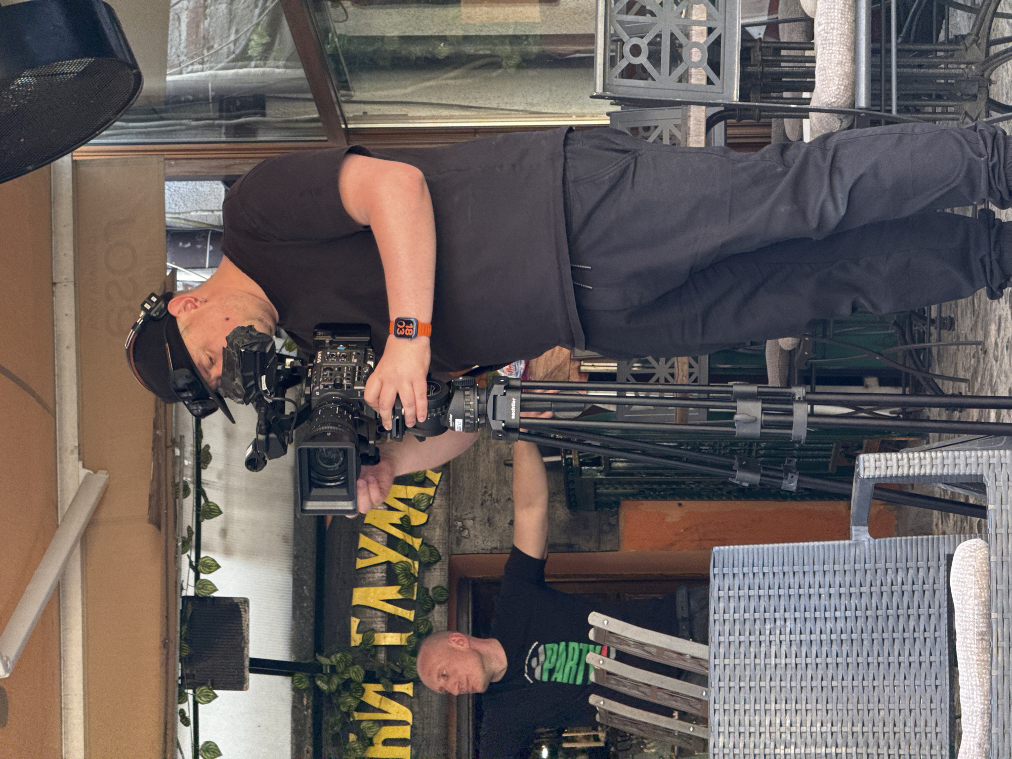 The image depicts a scene in what appears to be Belgrade, Serbia. A cameraman, wearing a black t-shirt, dark pants, and a baseball cap, is operating a professional video camera mounted on a tripod. He is looking through the viewfinder. Another person, wearing a black t-shirt with a graphic, is standing nearby. There is wicker furniture in the foreground. A sign with the word "LAZY" is visible in the background, along with some foliage. The environment suggests an outdoor setting, possibly a patio or terrace. A metal fan or light fixture is in the upper left.