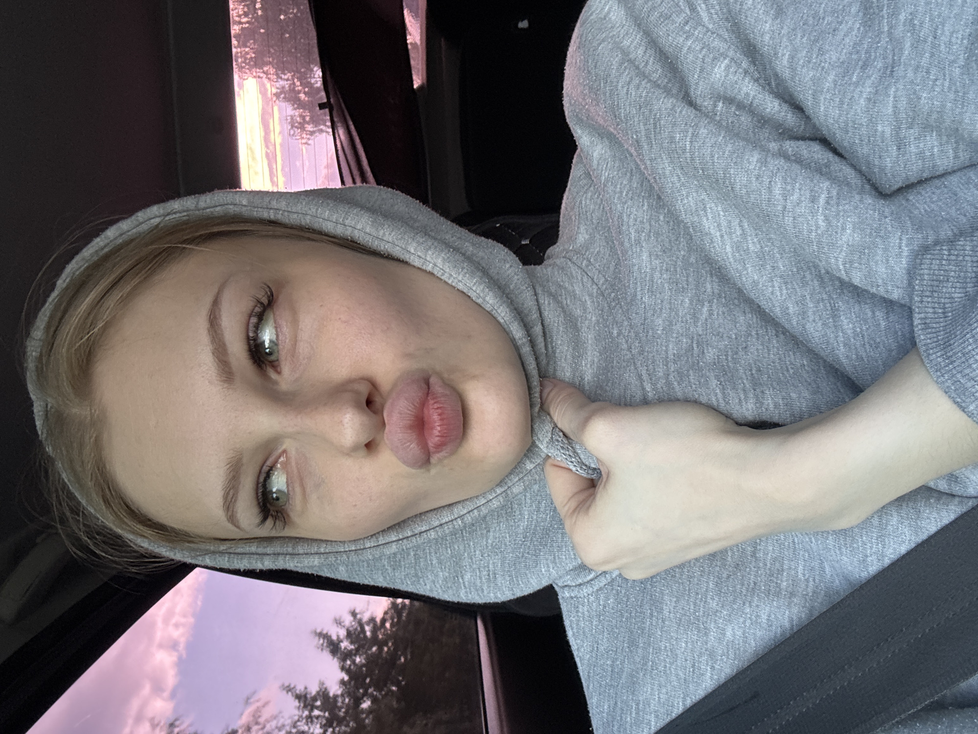 In the city of Perm, Russia, a young woman with blonde hair, wearing a grey hoodie, is pictured in what appears to be the interior of a vehicle. She is looking to her left with her lips pursed. The background shows trees and a sky with pinkish clouds, suggesting either sunrise or sunset. A seatbelt is visible across her shoulder.
