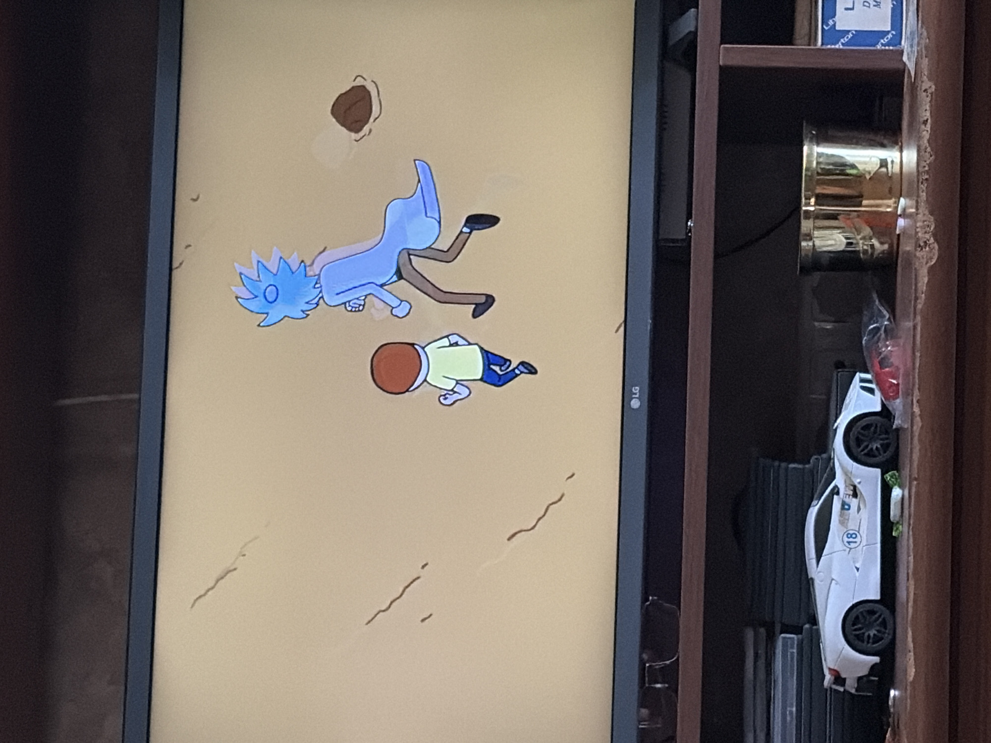 The image shows a television screen displaying a scene from the animated show Rick and Morty. The characters Rick Sanchez and Morty Smith are depicted running across a sandy landscape. Rick, with his distinctive blue spiky hair and lab coat, is in the foreground, followed by Morty, who has brown hair and is wearing a yellow shirt and blue pants. To the right of the characters, a brown, round object rests on the ground. The television is placed on a dark wooden shelf, above a collection of DVDs and a white toy sports car. To the right of the car, two gold-colored cylindrical containers are visible. The overall environment is dimly lit, suggesting an indoor setting.