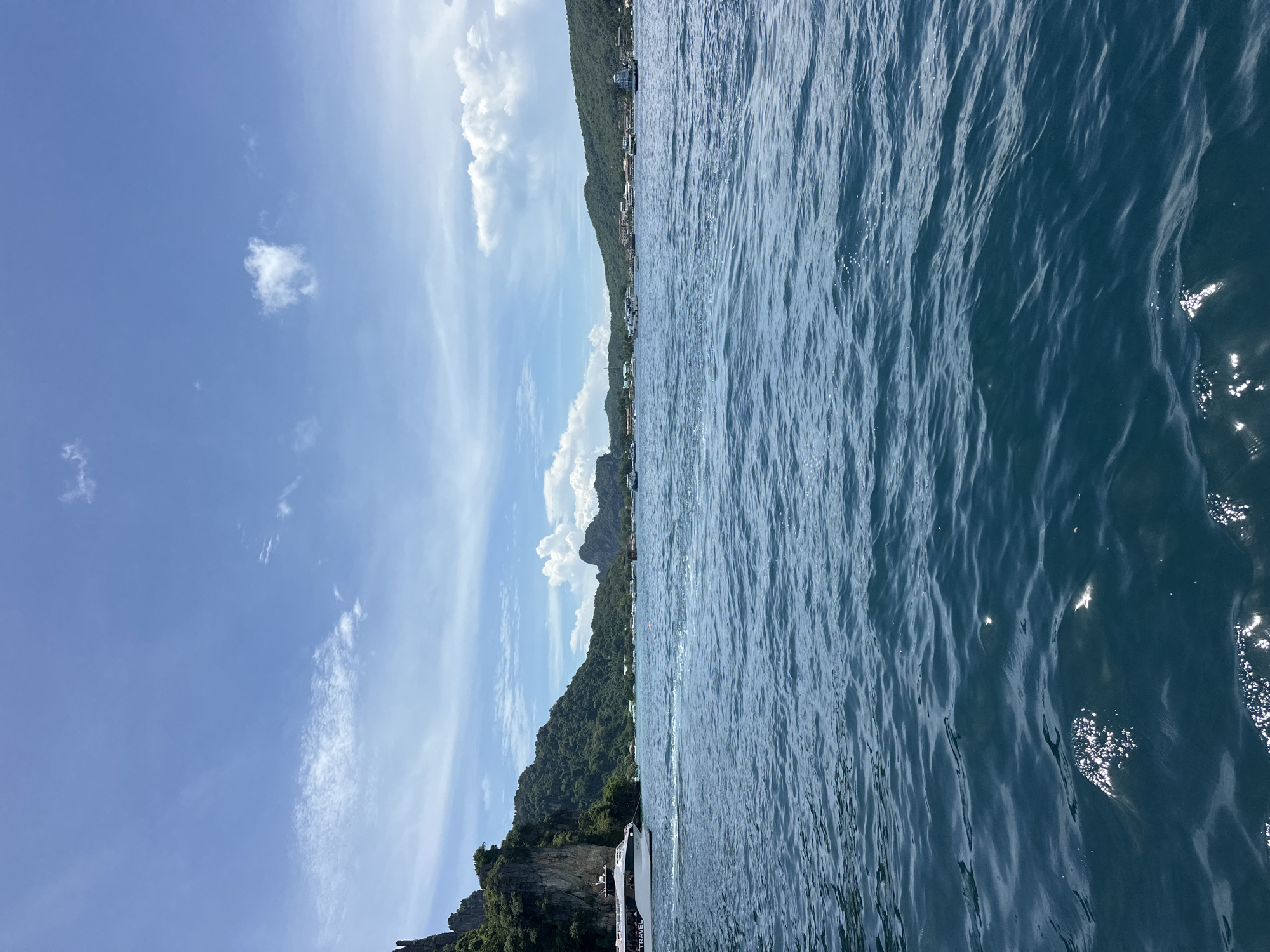 The image shows a view from a boat on the ocean near Ao Nang, Thailand. The water is a deep blue and has small waves. In the distance, there are several islands covered in lush green vegetation and rocky cliffs. The sky is blue with scattered white clouds. A large white ferry boat is visible on the left side of the image, moving towards the right. Other smaller boats are scattered across the water in the background, near the shorelines of the islands. Buildings and structures are visible along the coastlines of the islands, suggesting a populated area.