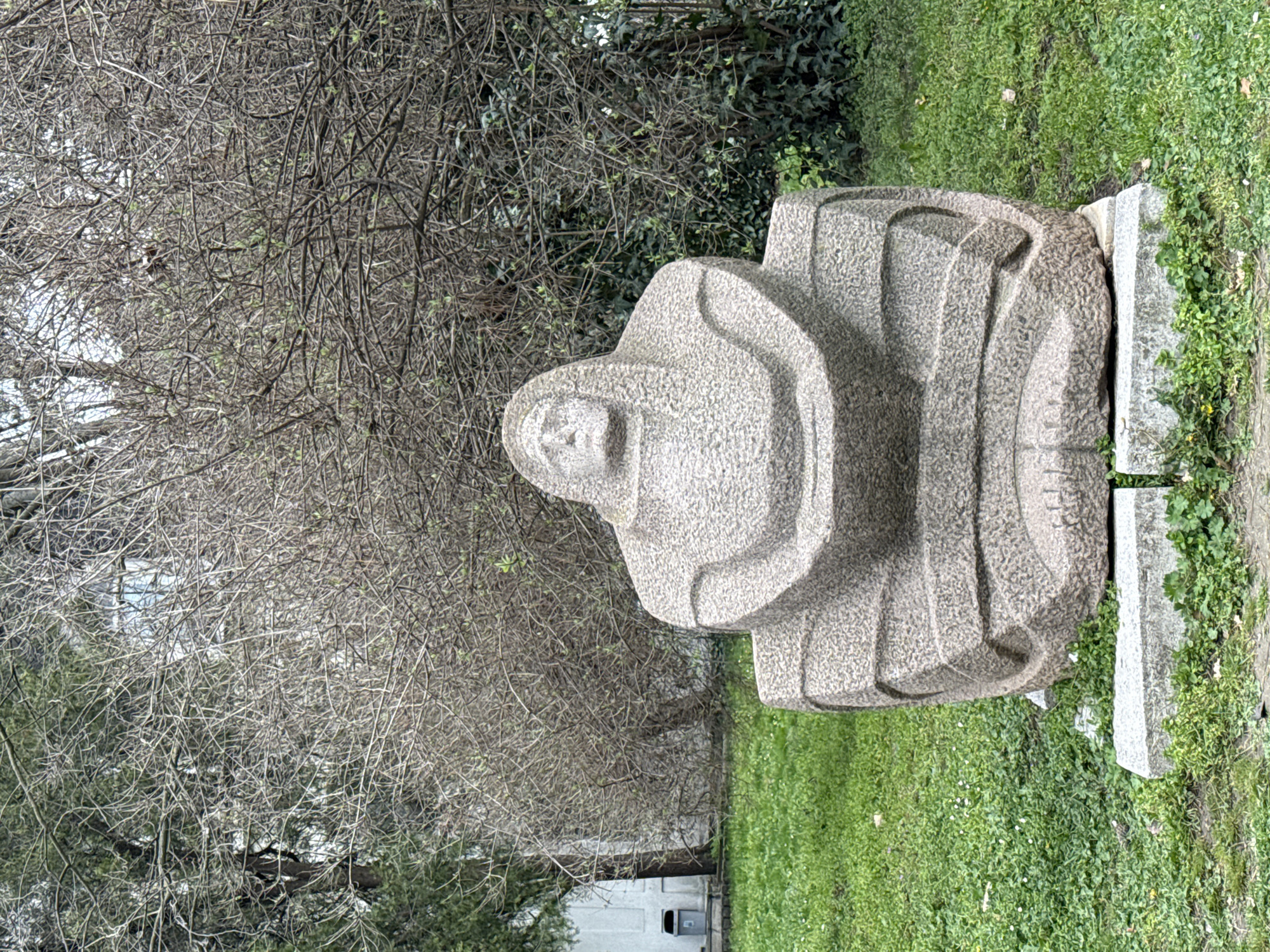 A medium shot depicts a stone sculpture of a robed figure located in Varna, Bulgaria. The sculpture is positioned on a grass lawn near a leafless tree. The sculpture rests on a concrete base composed of two blocks. A building is visible in the background.