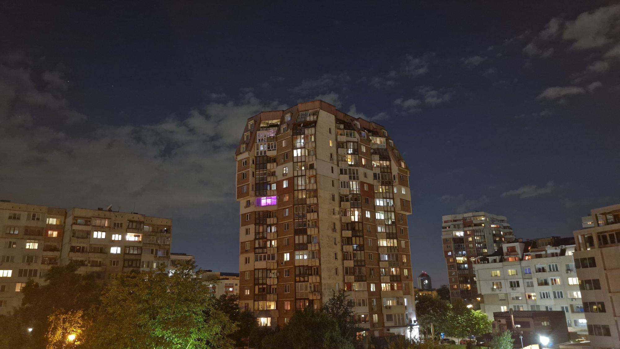 The image depicts an urban nighttime scene with a focus on apartment buildings. The main subject is a tall, prominent apartment building with numerous lit windows, suggesting occupancy. The building has a unique, multi-tiered top. Other buildings of varying heights surround it, also with illuminated windows. Trees are visible in the foreground, partially obscuring the lower portions of the buildings. The sky is dark with scattered clouds. The location is Unknown, Unknown.