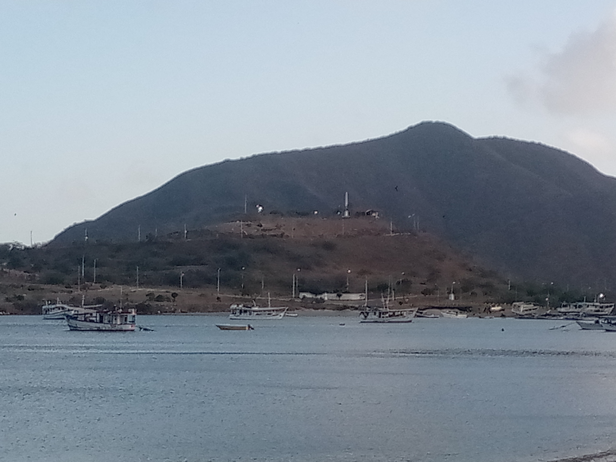 The image depicts a tranquil bay with several boats anchored in the calm blue water. In the background, a large, dark mountain range dominates the landscape, with a few scattered structures visible on its slopes. The sky is a pale blue, with a hint of clouds in the upper right corner. The overall scene suggests a peaceful, coastal environment, possibly in a location with limited development.