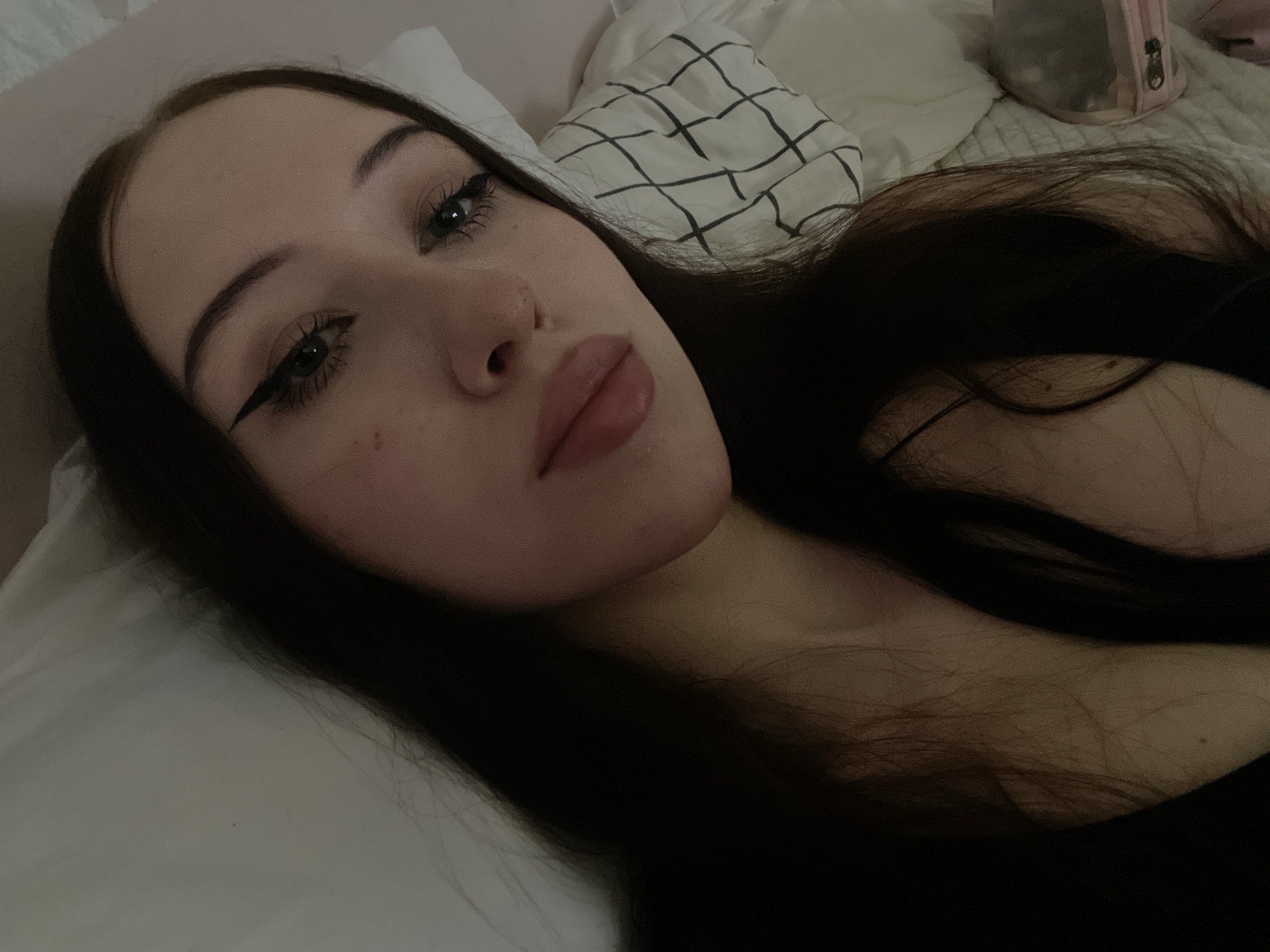 The image shows a close-up, high-angle, indoor, selfie-style photograph of a young woman with long, dark hair. She is lying down, with her head resting on a pillow patterned with a black grid. The woman is wearing makeup, including eyeliner. Her skin is fair, and she has a light blush on her cheeks. She is wearing a dark-colored top, and her hair is spread around her. The background appears to be a bed with white bedding. There is no information available to determine the location in Votkinsk, Russia.