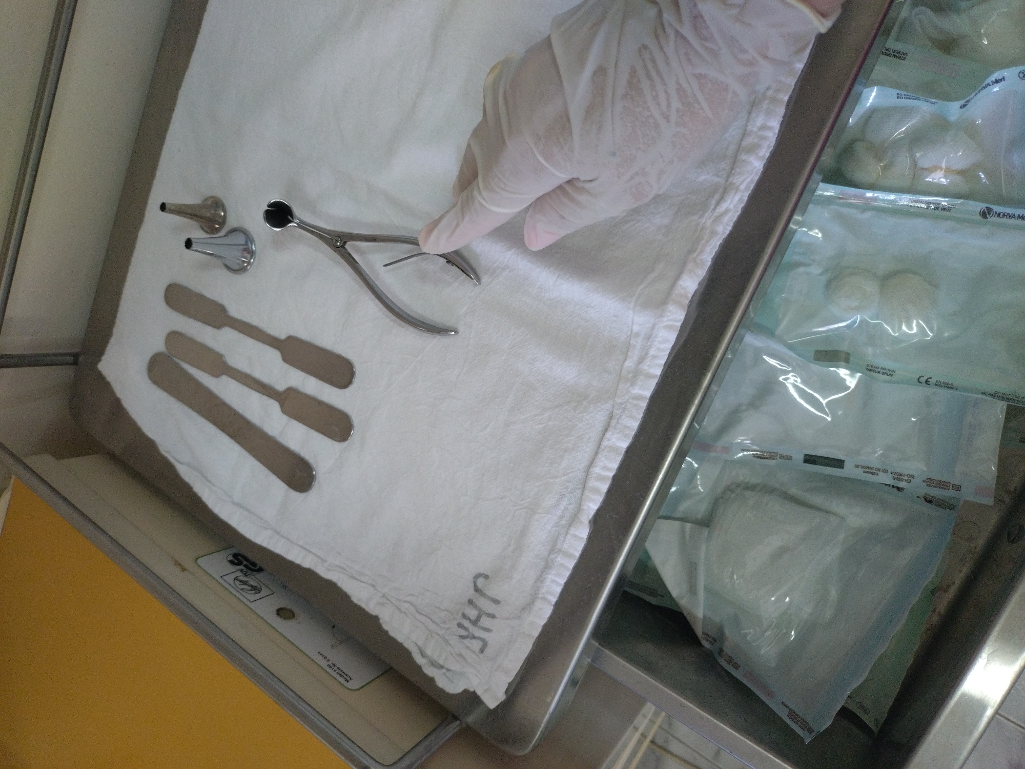 A gloved hand reaches for a pair of nasal forceps among other ENT medical instruments on a tray covered with a white cloth in what appears to be a medical setting. The tray is positioned next to a stainless steel drawer containing sealed packages of medical supplies. Visible text on the packages includes "STEAM BROWN", "VAPEUR BRUN", "NORVA Medi", "Auswels-N", and "Med". The drawer and tray are set on a yellow surface. The location is undetermined.