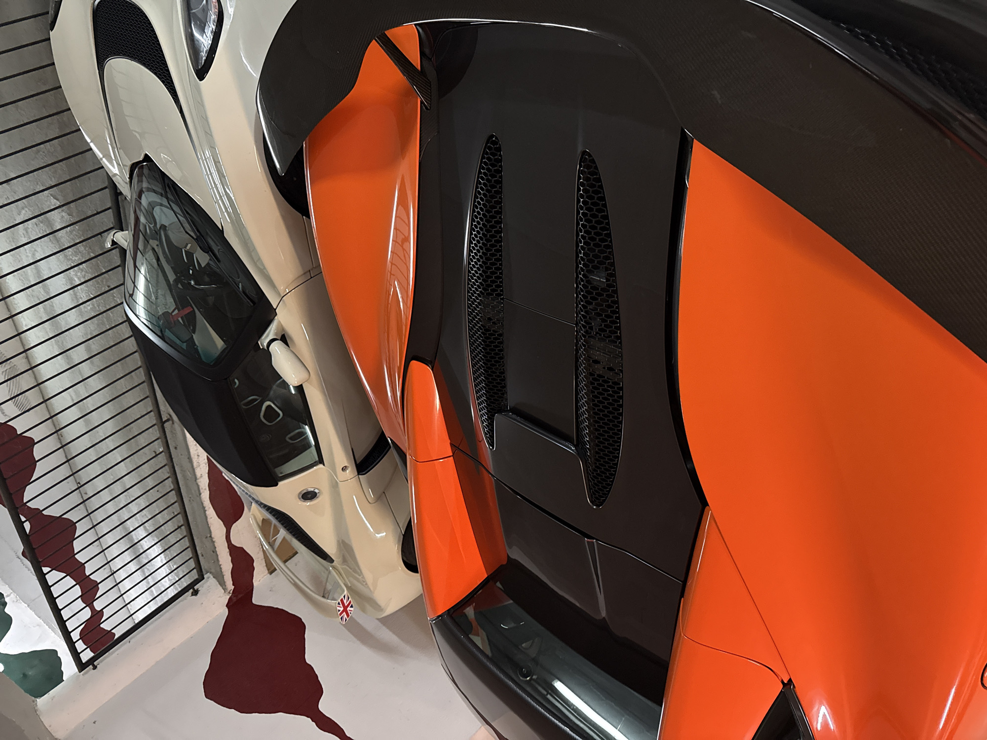 The image depicts the rear end of two luxury sports cars parked in what appears to be a garage or showroom. The car on the right is predominantly orange and black, featuring carbon fiber accents and visible air vents. To its left is a cream-colored car with black trim, with a Union Jack sticker near its front. The floor of the garage is painted white with irregularly shaped red and green blotches. A black metal railing is partially visible in the lower left corner of the image. The location, Benasque, Spain, is not explicitly evident within the image itself.