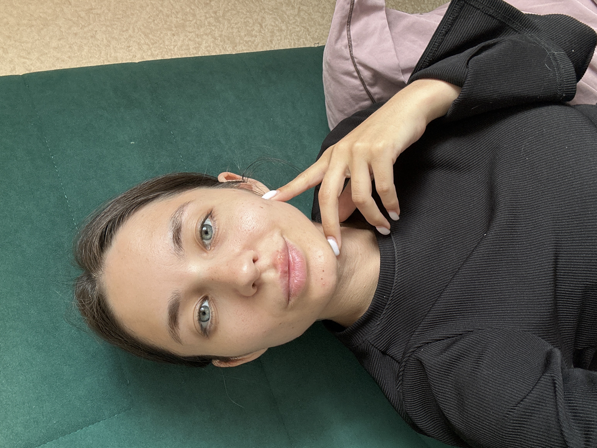 The media file is a selfie of a young woman lying on a dark green surface, possibly a couch or bed. She is looking directly at the camera with a neutral expression. Her head is tilted, and her right hand is raised, with her index finger touching her cheek near her eye. She is wearing a black, ribbed long-sleeved top. A pale pink pillow is visible behind her. The lighting suggests it is daytime, likely indoors. There are no visible text elements in the image. The setting appears to be a residential or casual environment.