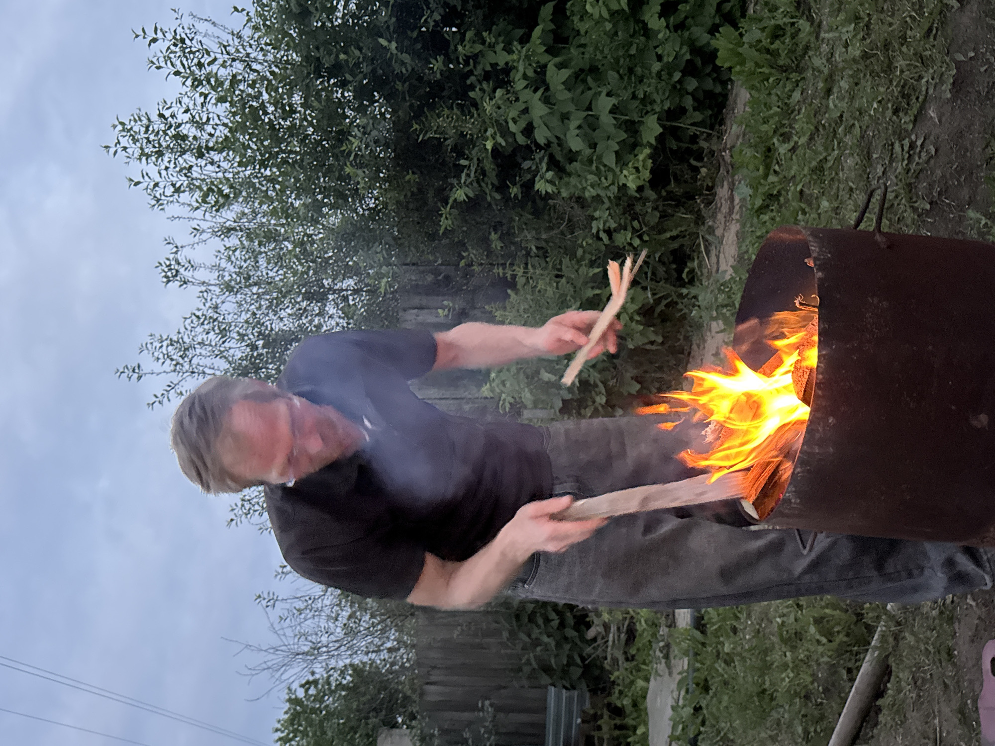 A man with blond hair and glasses is tending a fire in a metal barrel in an outdoor setting that appears to be a backyard in Chaikovsky, Russia. He is wearing a dark t-shirt and grey pants. The man is holding a wooden stick in his right hand and is using it to adjust the burning wood in the barrel. Flames are visible in the barrel, and smoke is rising. The background consists of lush green foliage and a wooden fence, suggesting a natural or semi-rural environment. The sky is overcast and appears to be dusk.