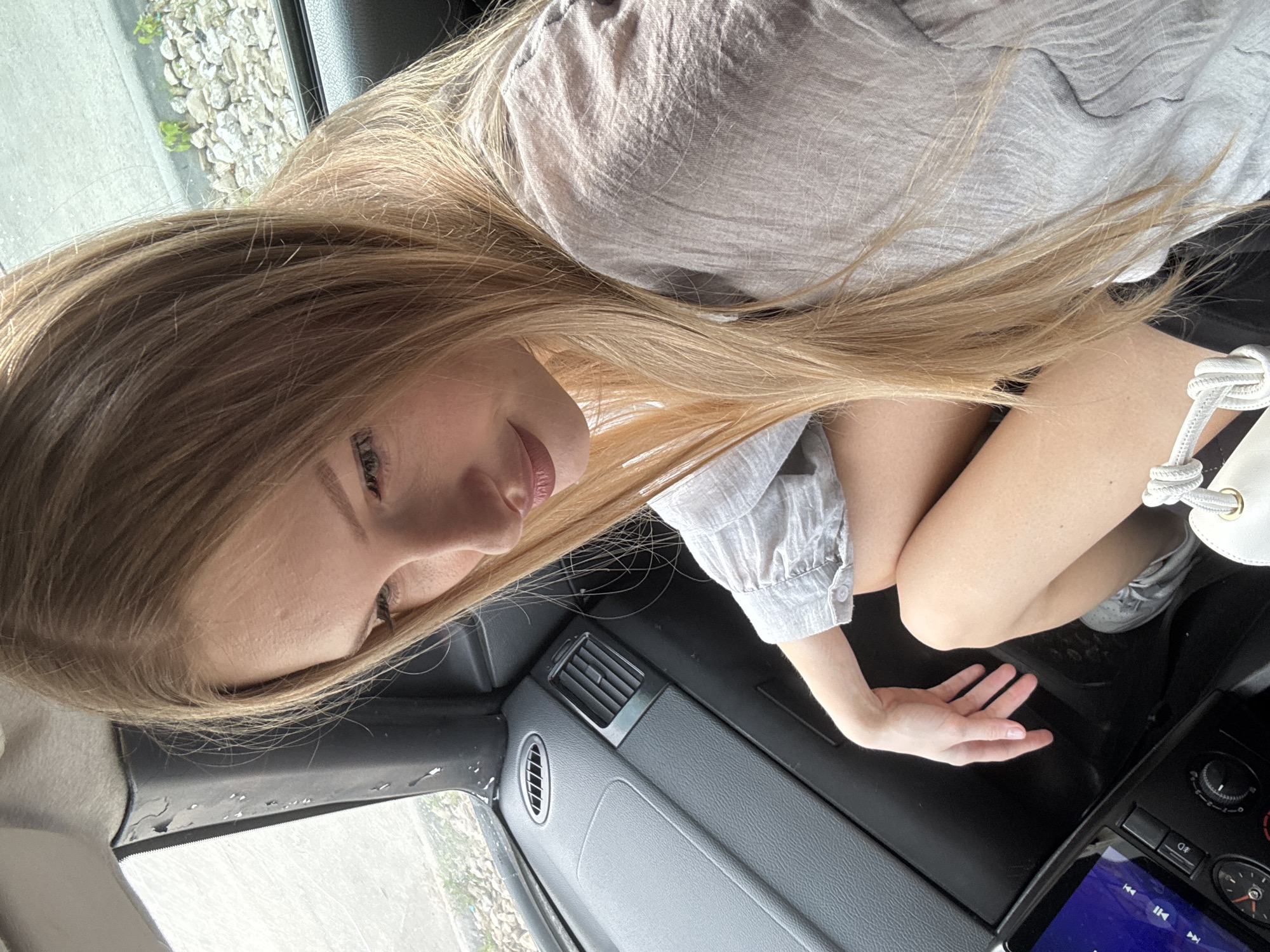 A young woman with blonde hair is sitting in the passenger seat of a car. She is looking down and to the side. Her right arm is resting on her leg, and her left hand is on the car's dashboard. She is wearing a gray shirt and shorts. The car's interior is visible, including the dashboard, air vents, and a touchscreen display. The exterior of the car is visible through the window, showing a street with some gravel and greenery. The weather appears to be overcast. The image was taken in Perm, Russia.