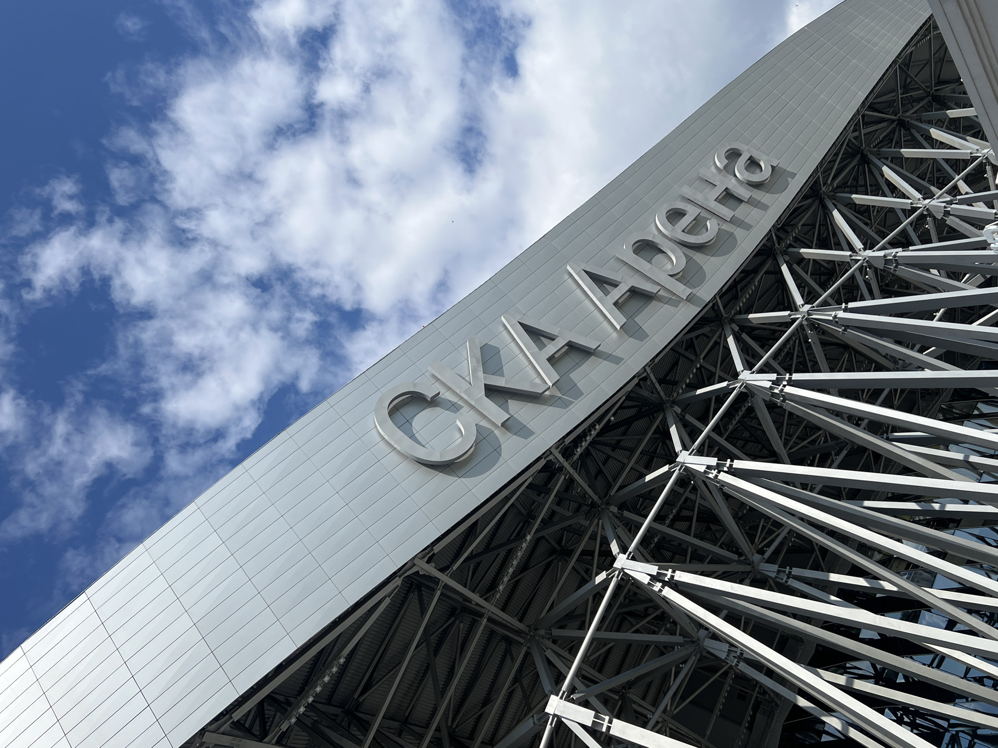 The image shows a low-angle view of the exterior of the SKA Arena in Saint Petersburg, Russia. The building features a modern, metallic facade and a complex, exposed steel support structure. Large, three-dimensional letters spelling out "SKA Арена" are mounted on the facade. The sky above is blue with scattered white clouds.